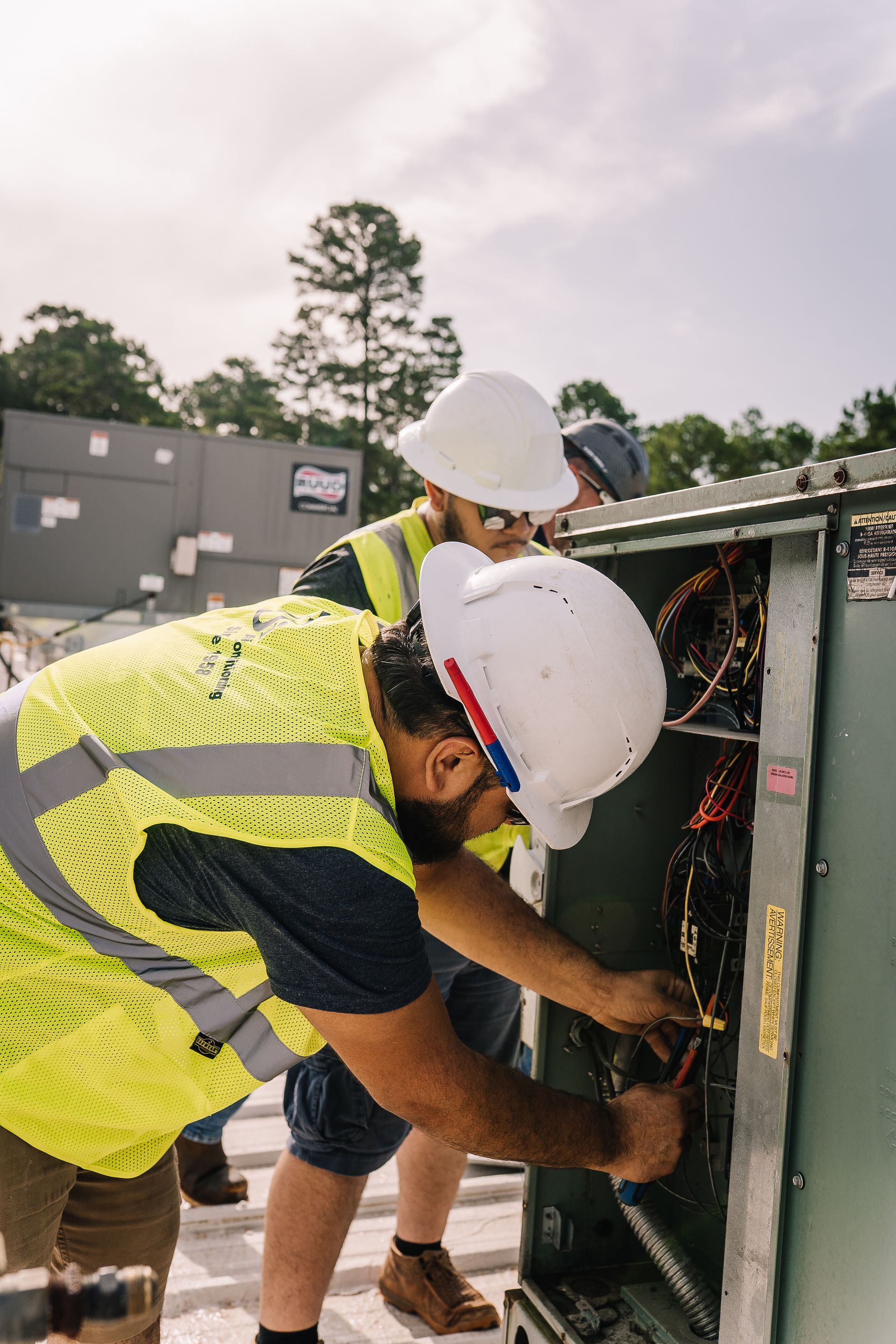 Two electricians in safety vests and hard hats work on wiring outdoors.