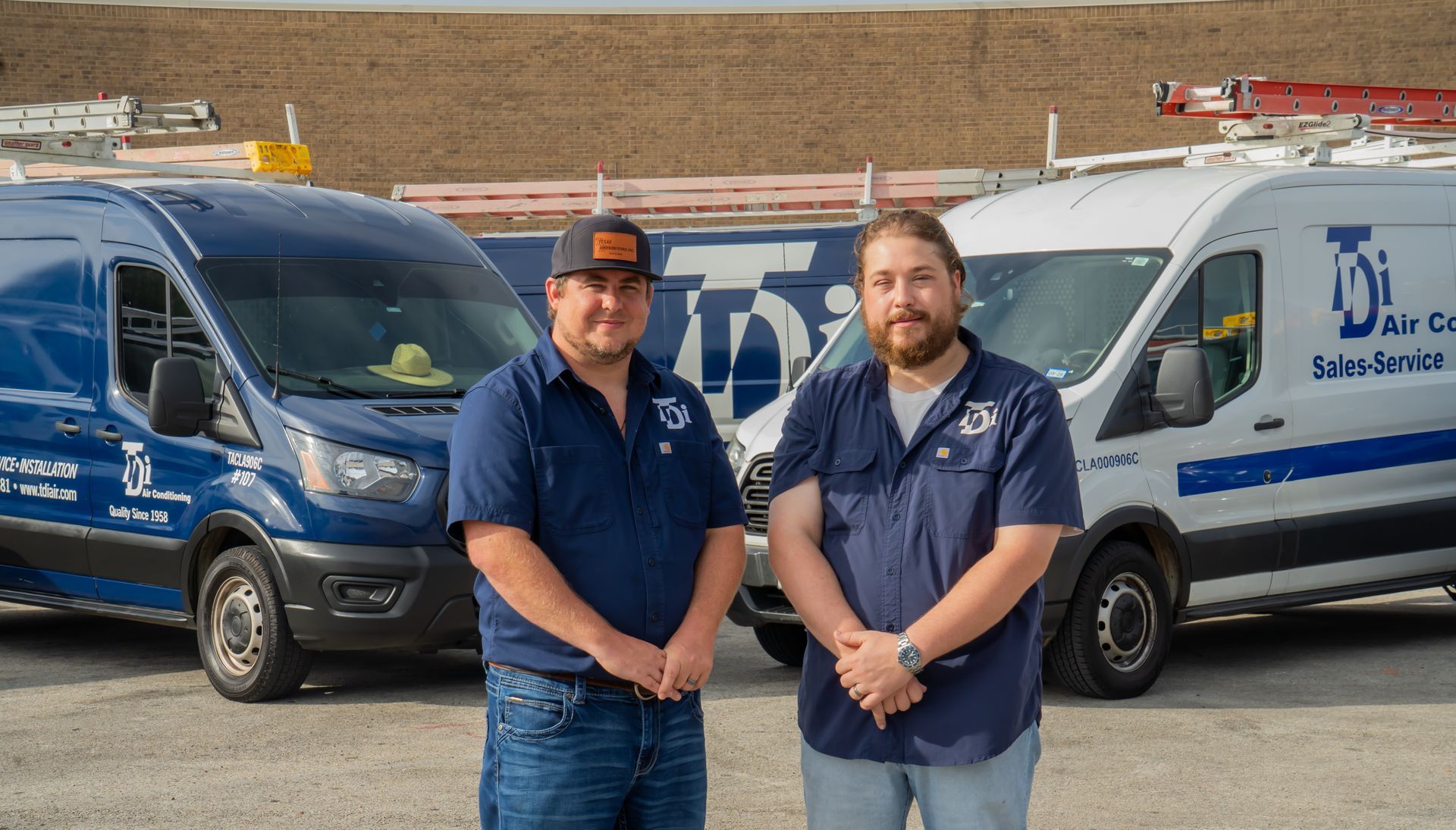 Two men in blue work shirts stand in front of work vans.