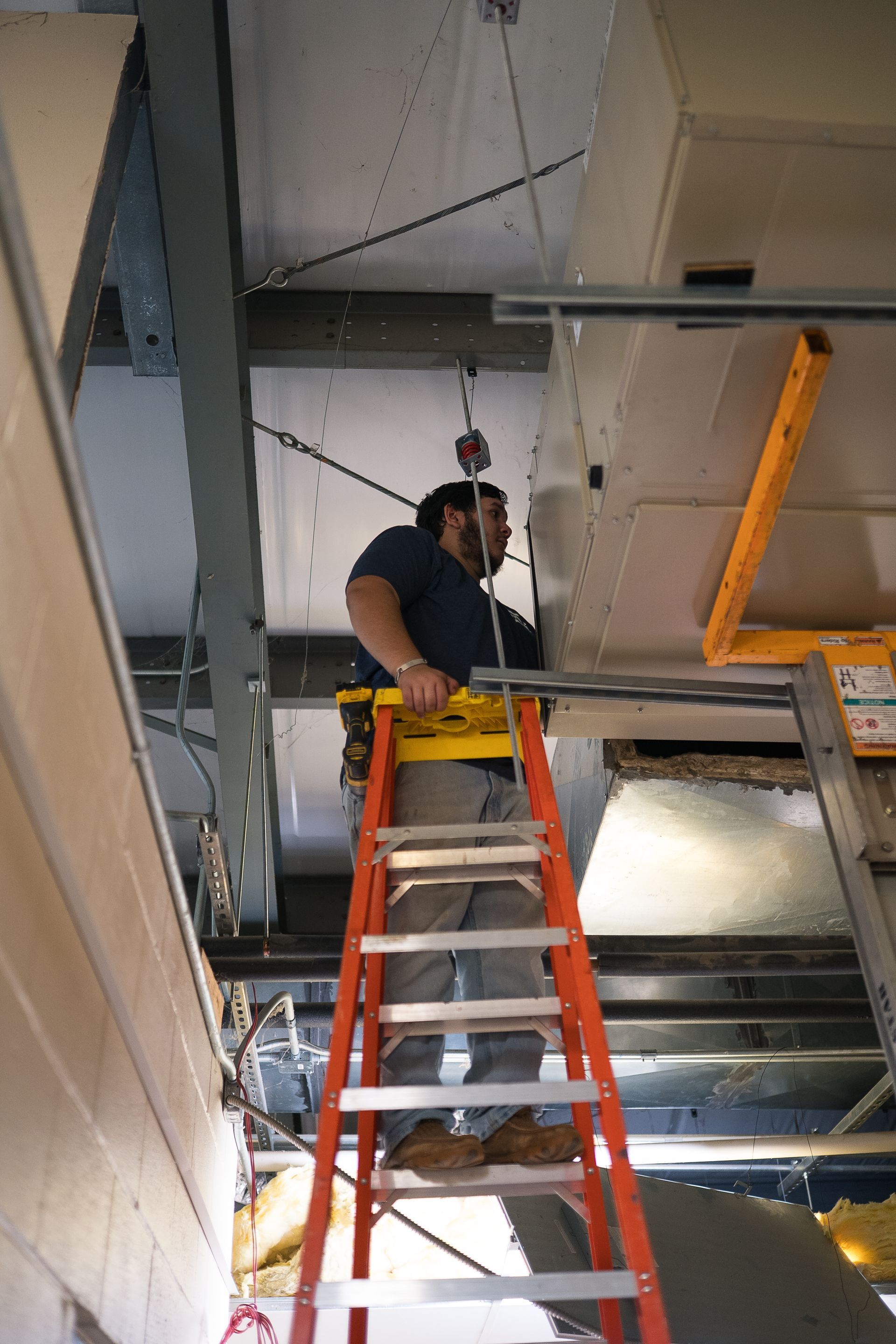 Man on orange ladder, installing something on ceiling, near ductwork.