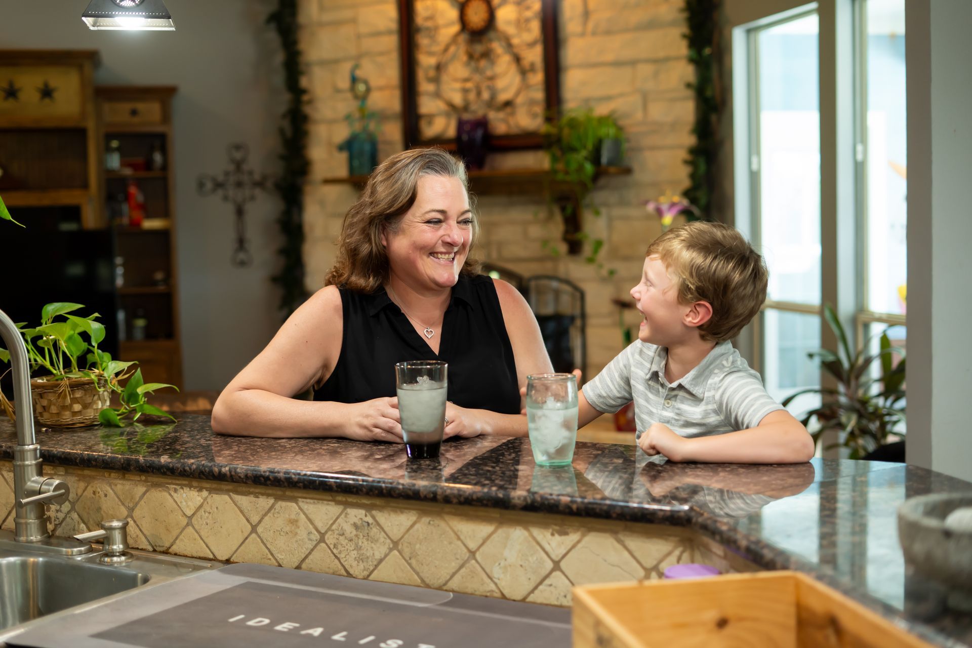 Woman and boy smiling at each other over a kitchen counter, each holding a drink with ice.