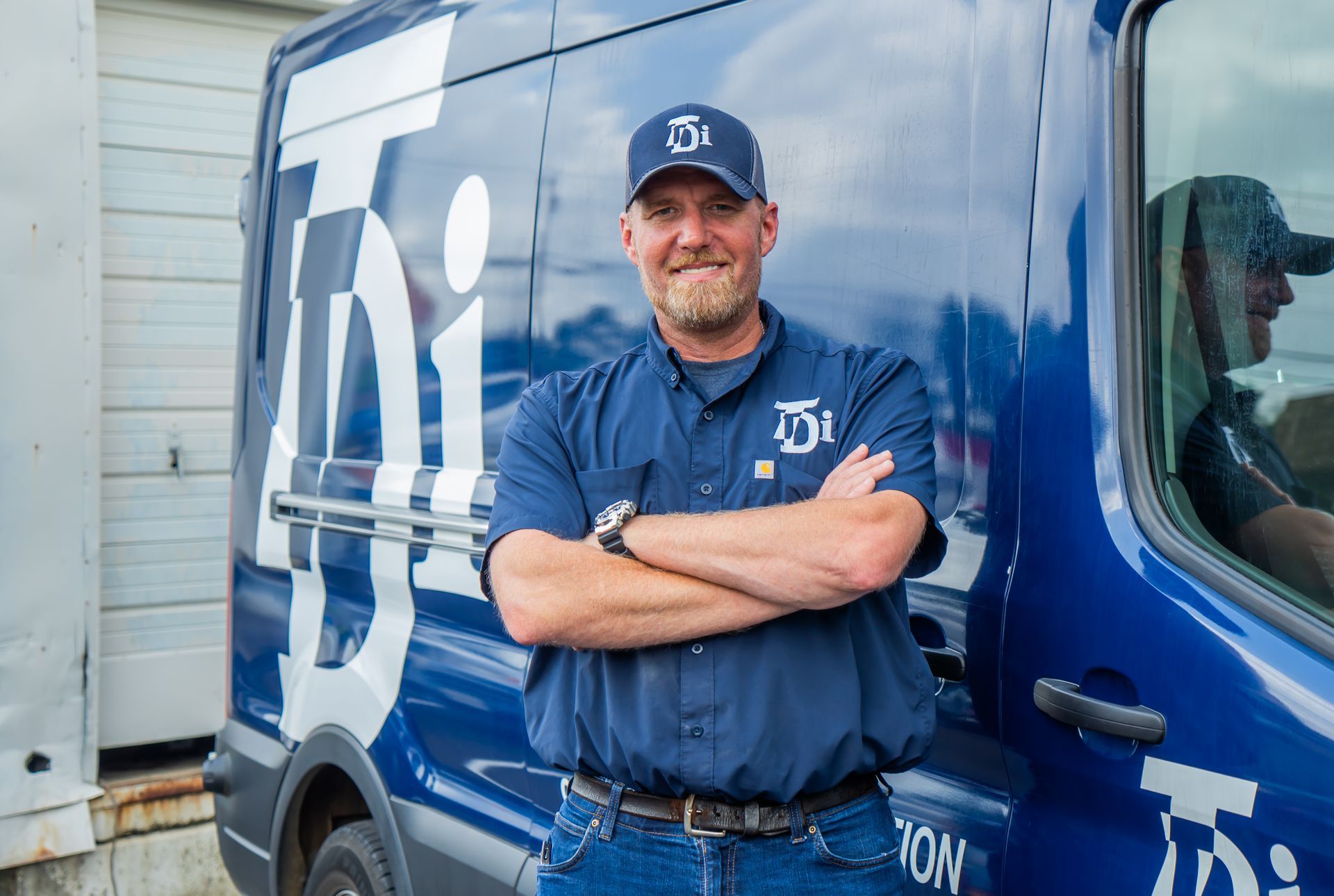 Man in work uniform, arms crossed, smiles in front of blue van with company logo.