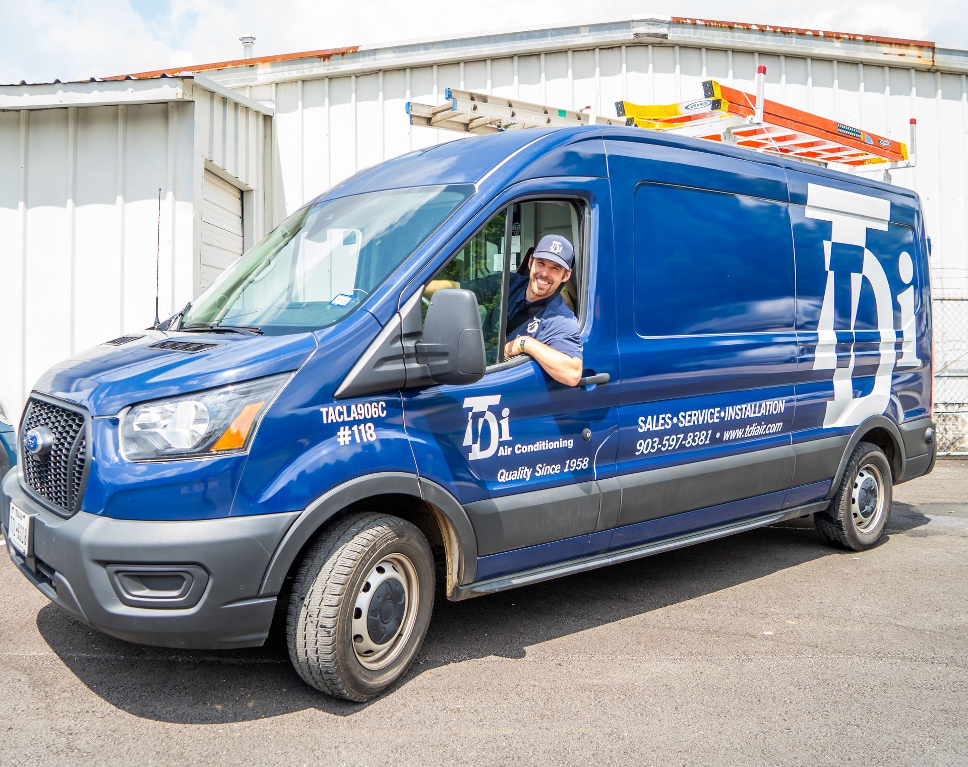 Blue service van with a smiling man in the driver's seat. Building in background.