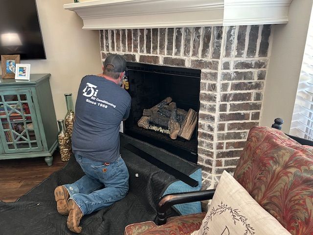 A man kneeling inside a fireplace, inspecting the gas logs. He wears a blue shirt and jeans. Brick surround.