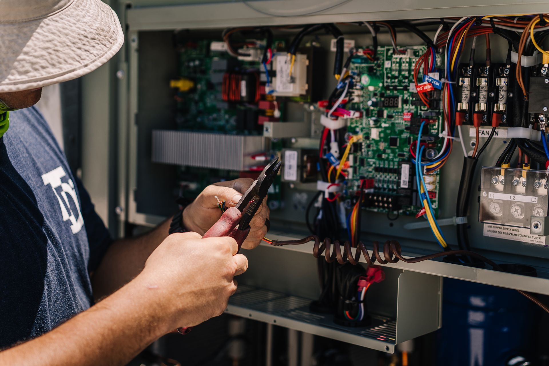 Person in a hat working on an electrical panel outdoors with tools.