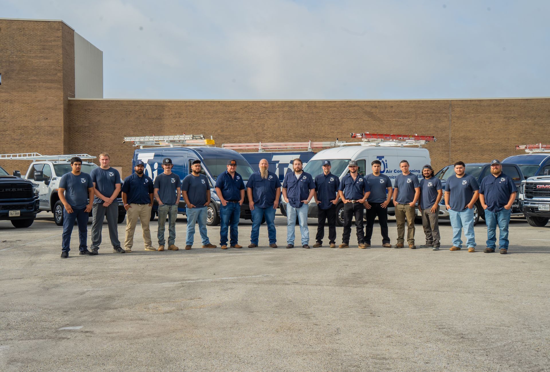 Group of men in blue shirts posing in front of company vans. Building in the background.