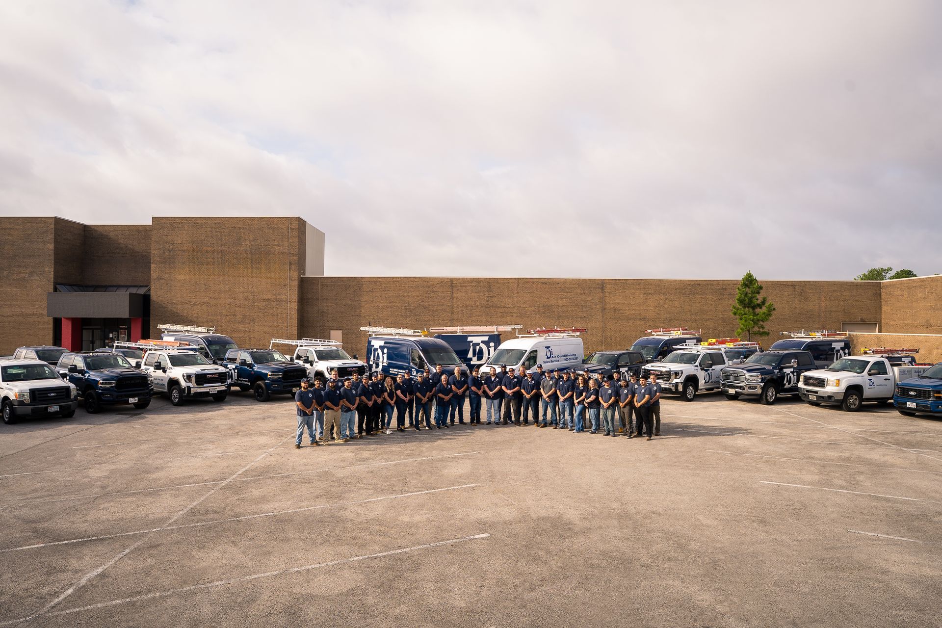Group of people with trucks in a parking lot in front of a brick building under a cloudy sky.