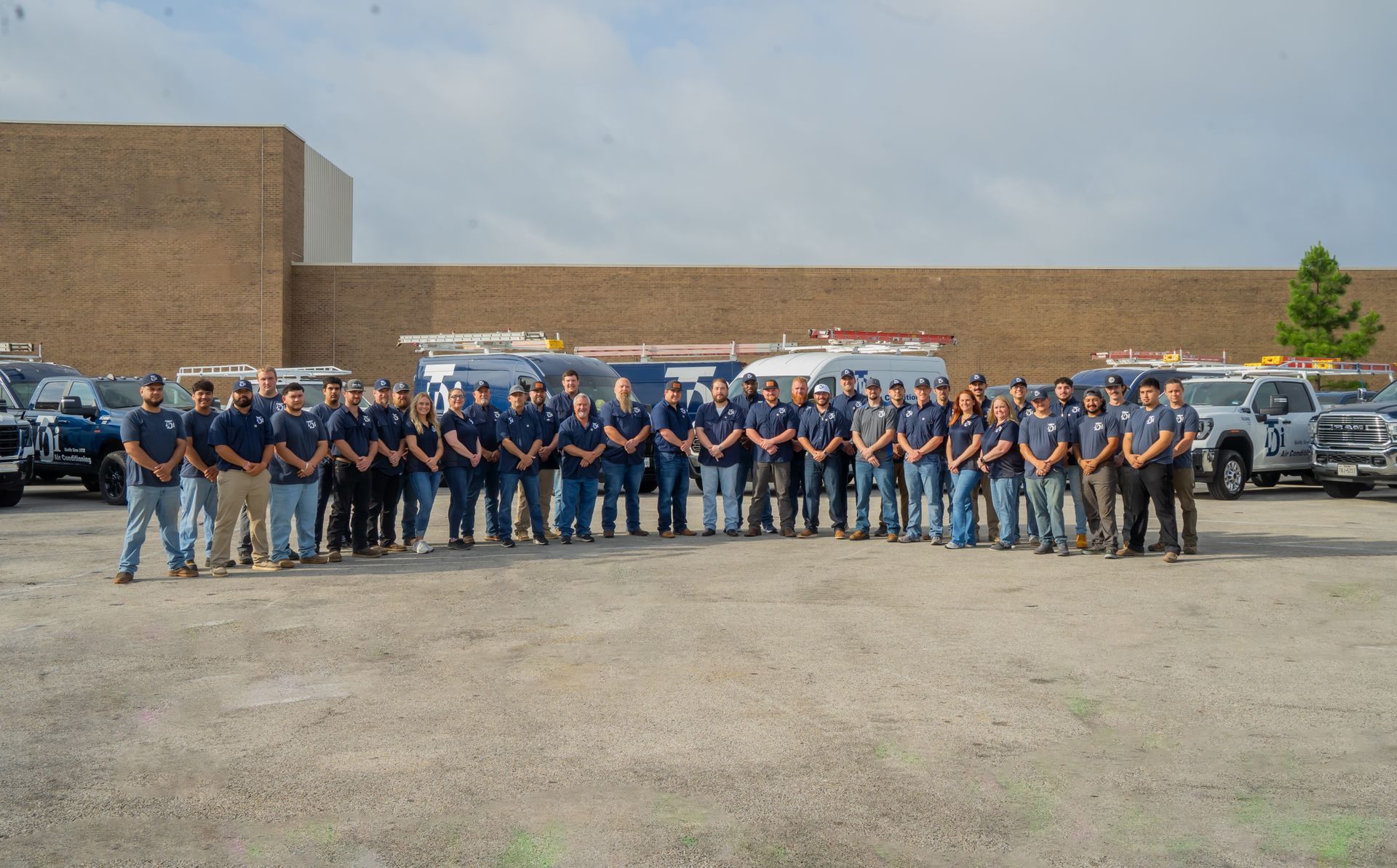 Group of workers in blue shirts and jeans, standing outdoors near trucks, in front of a brick building.
