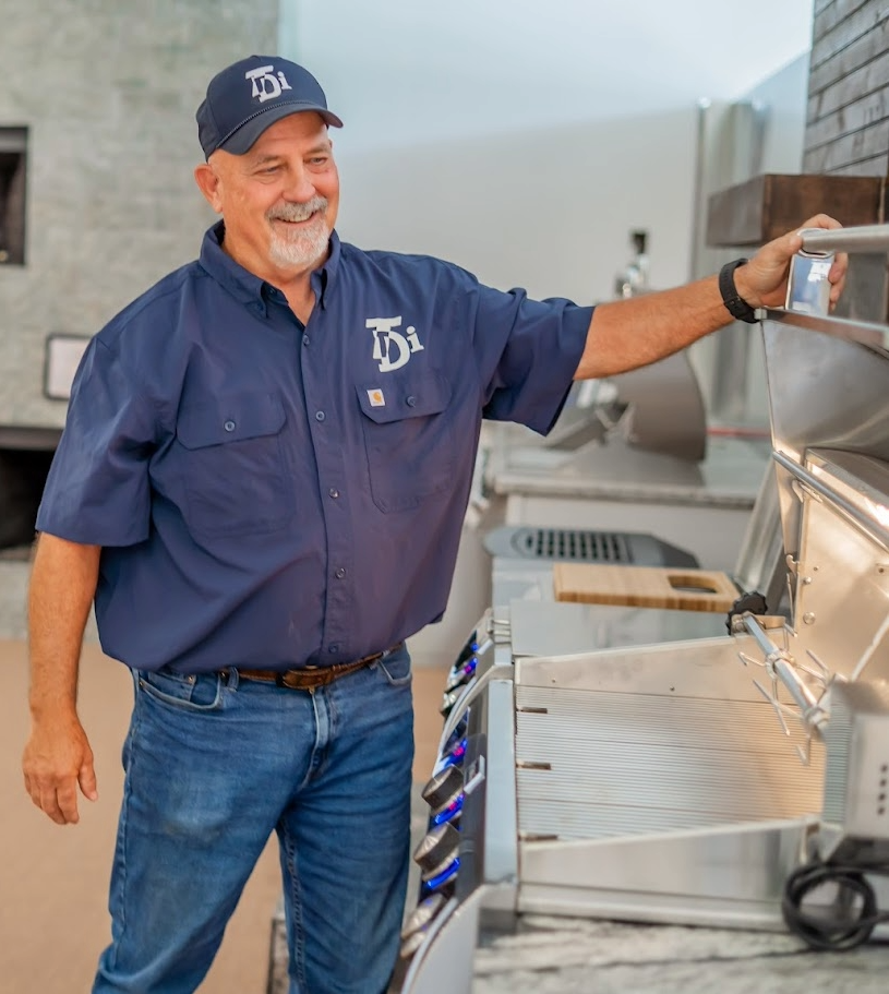 Man in blue shirt and jeans, smiling, looking at a grill. He wears a cap, in a modern kitchen.
