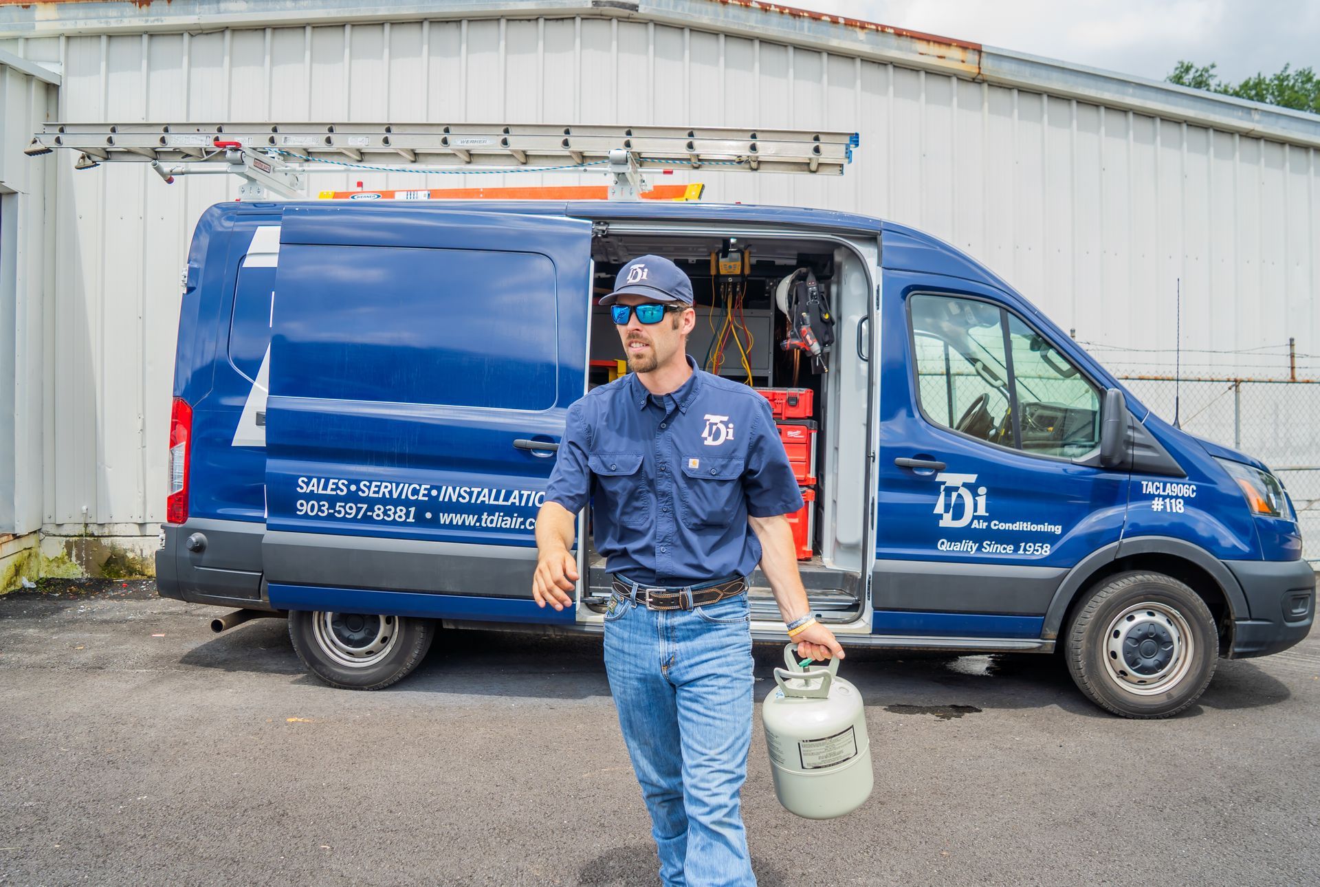 Man in uniform holding propane tank, exiting blue work van. Building in background.