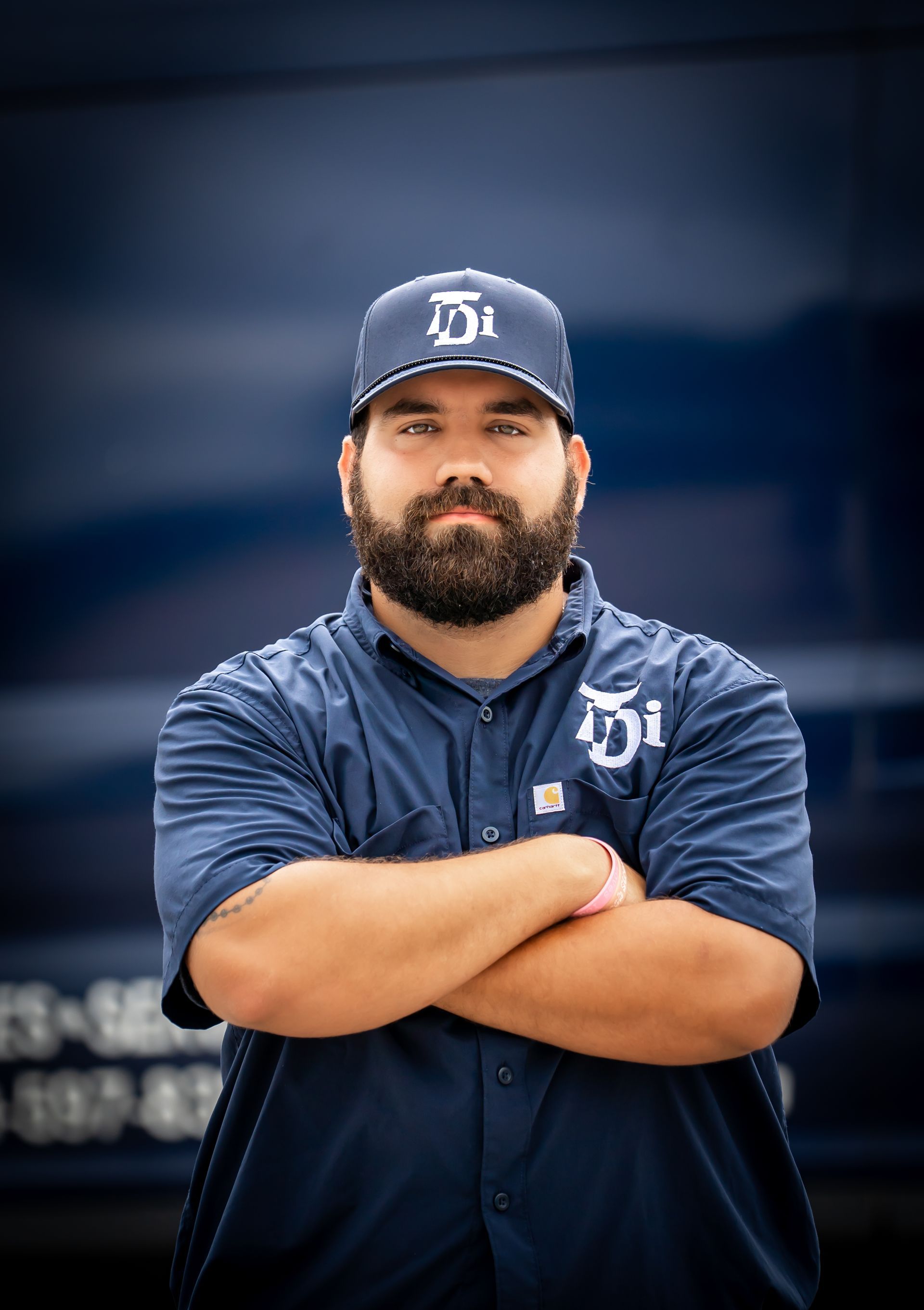 Man with beard, wearing a blue uniform and cap with logo, arms crossed, standing in front of a blue truck.