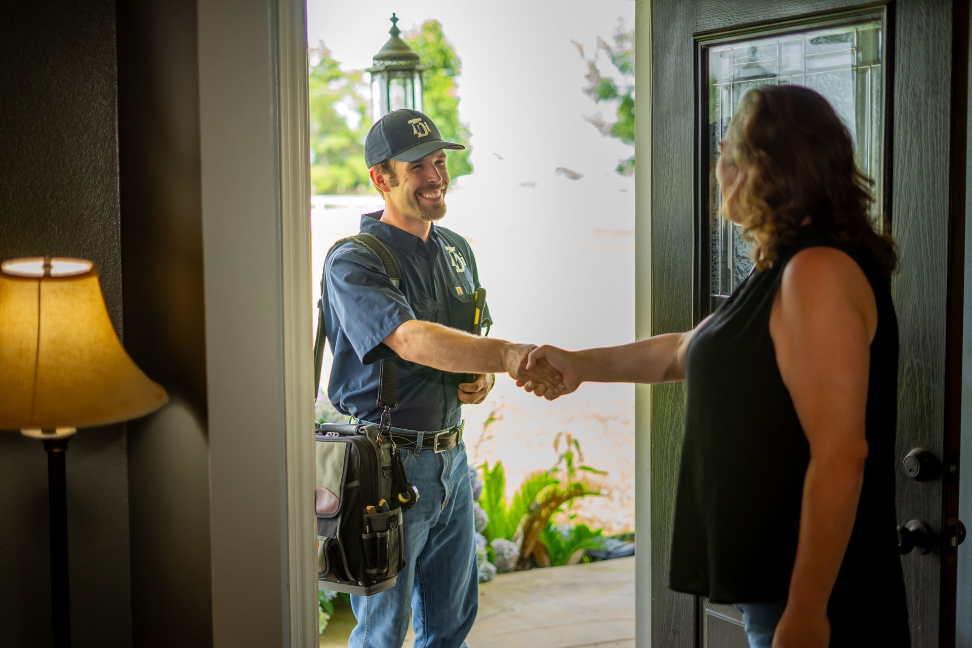 A smiling worker shakes hands with a woman at a doorway. The worker wears a uniform and tool belt.