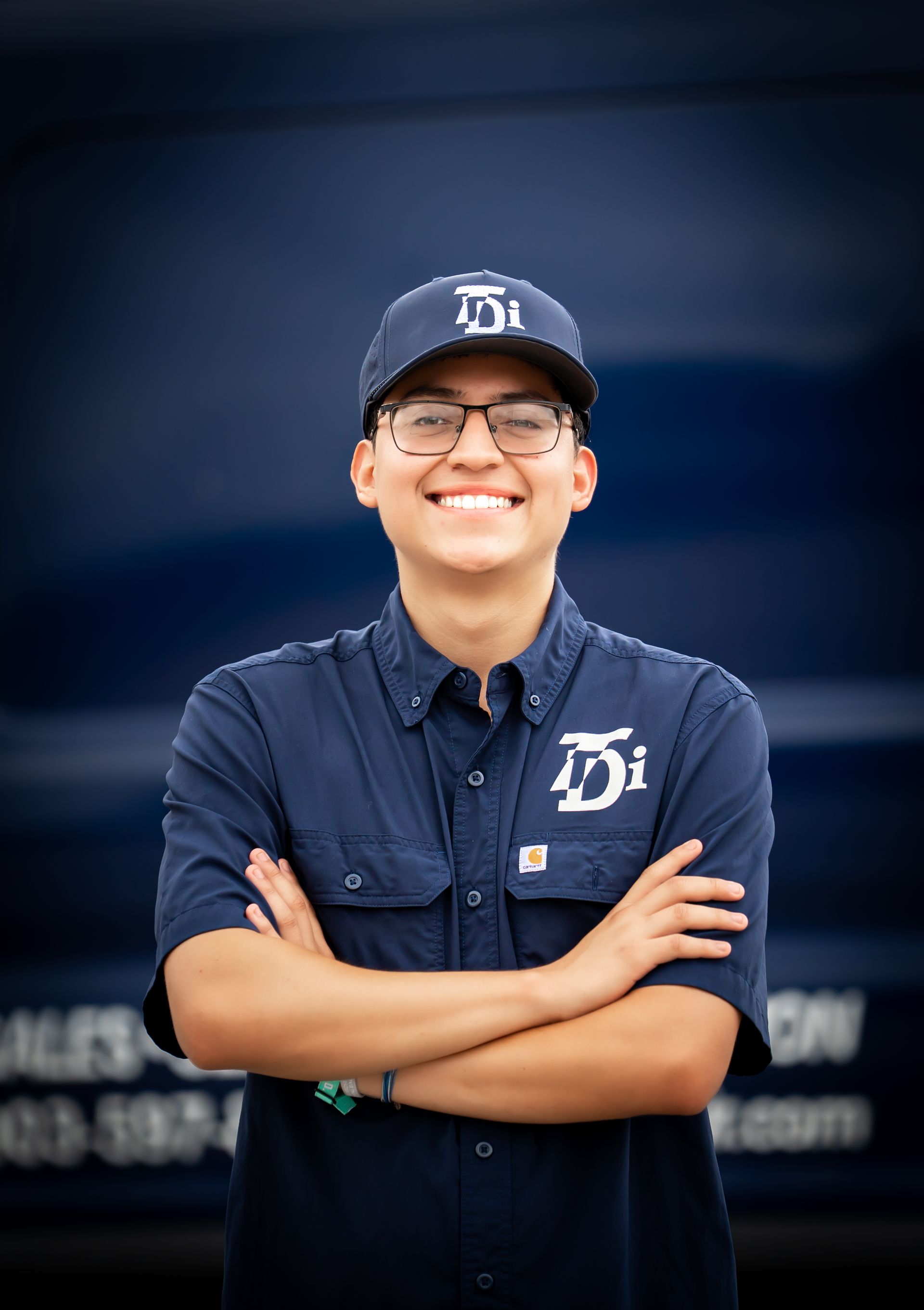 Young person in a blue uniform with crossed arms, wearing a hat and glasses, smiling in front of a blue vehicle.
