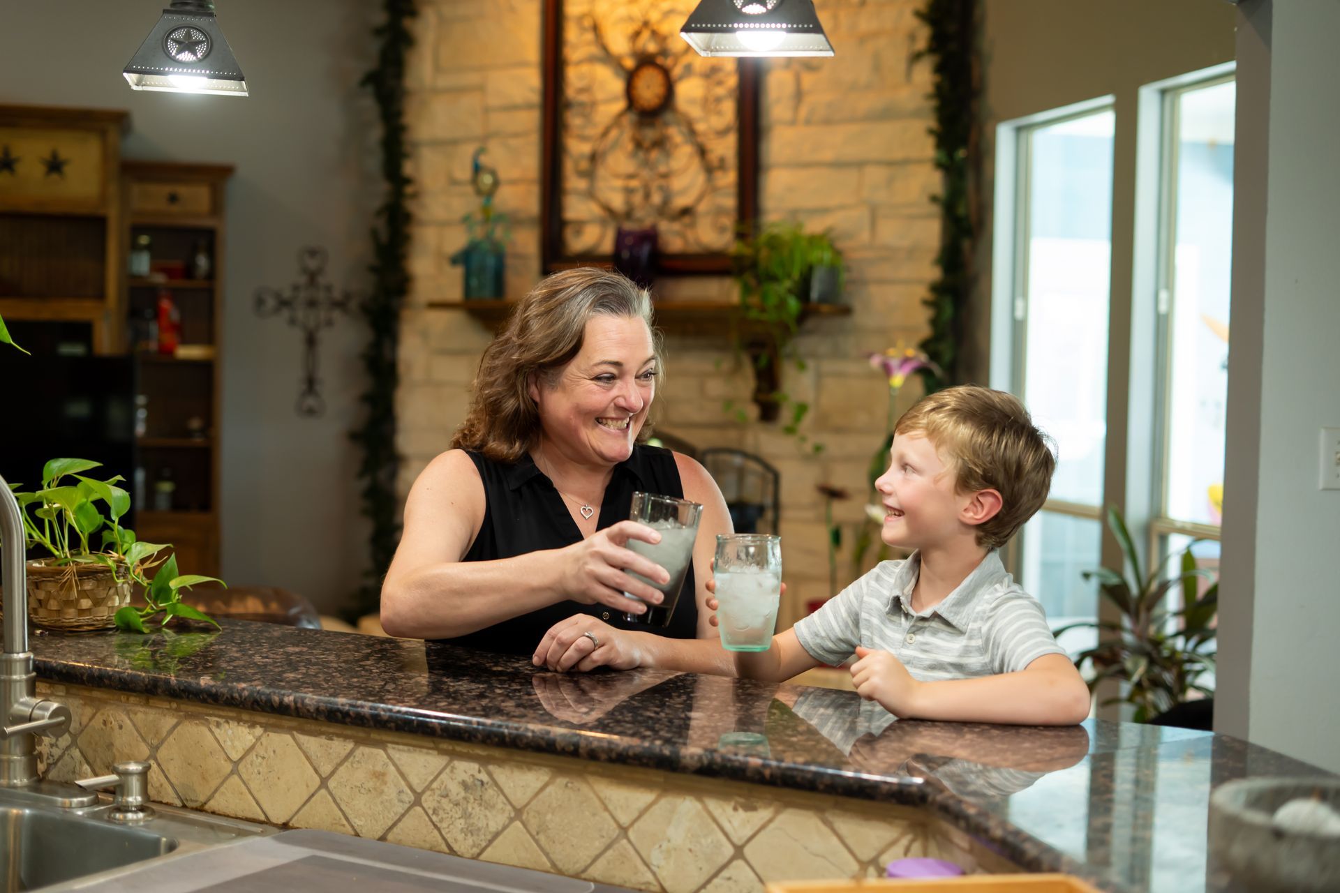 Woman and child, smiling, clink glasses filled with a white drink at a kitchen counter.