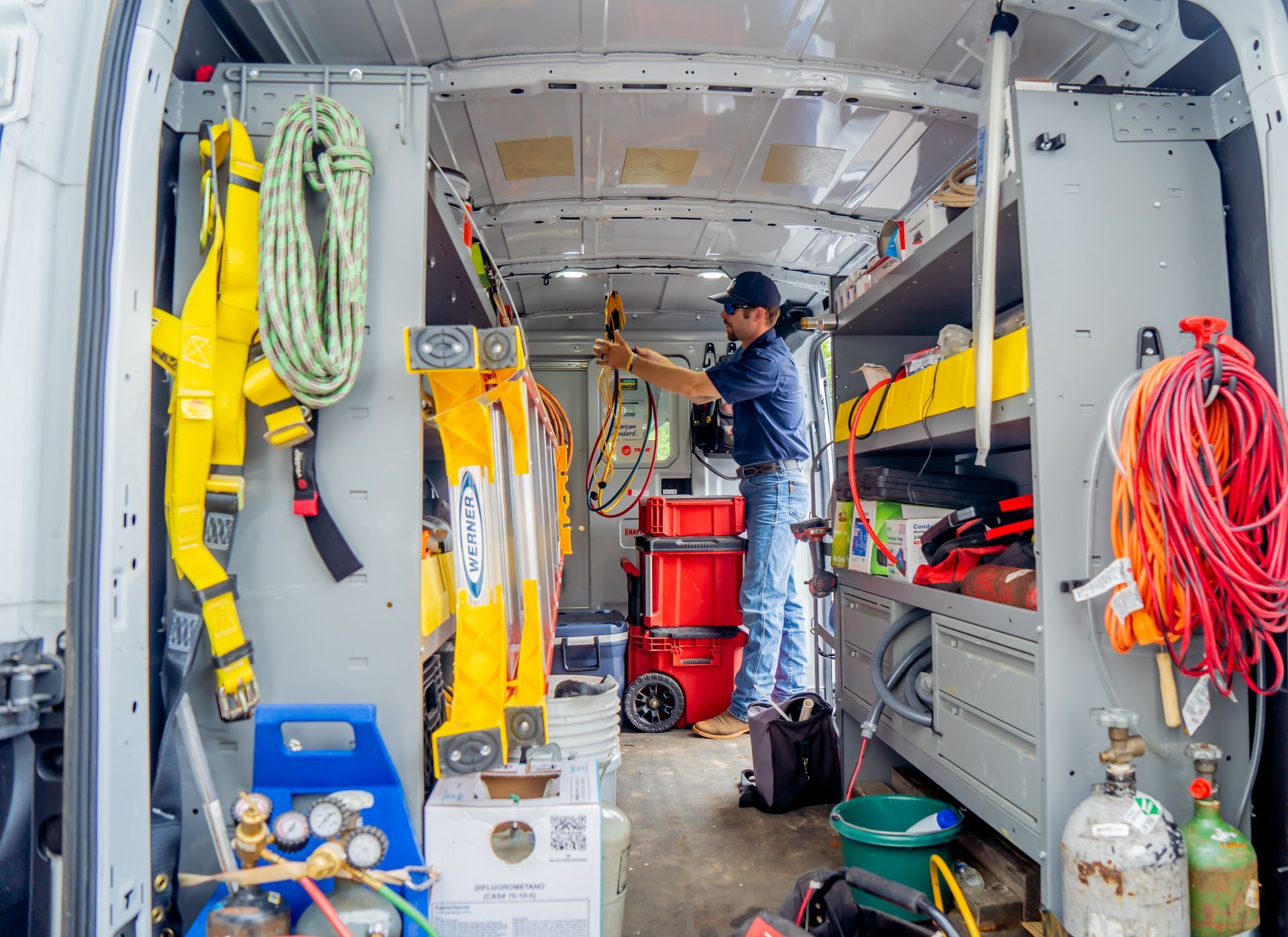 Man inside a work van organizing tools. Yellow ladder and ropes. Shelves with supplies.