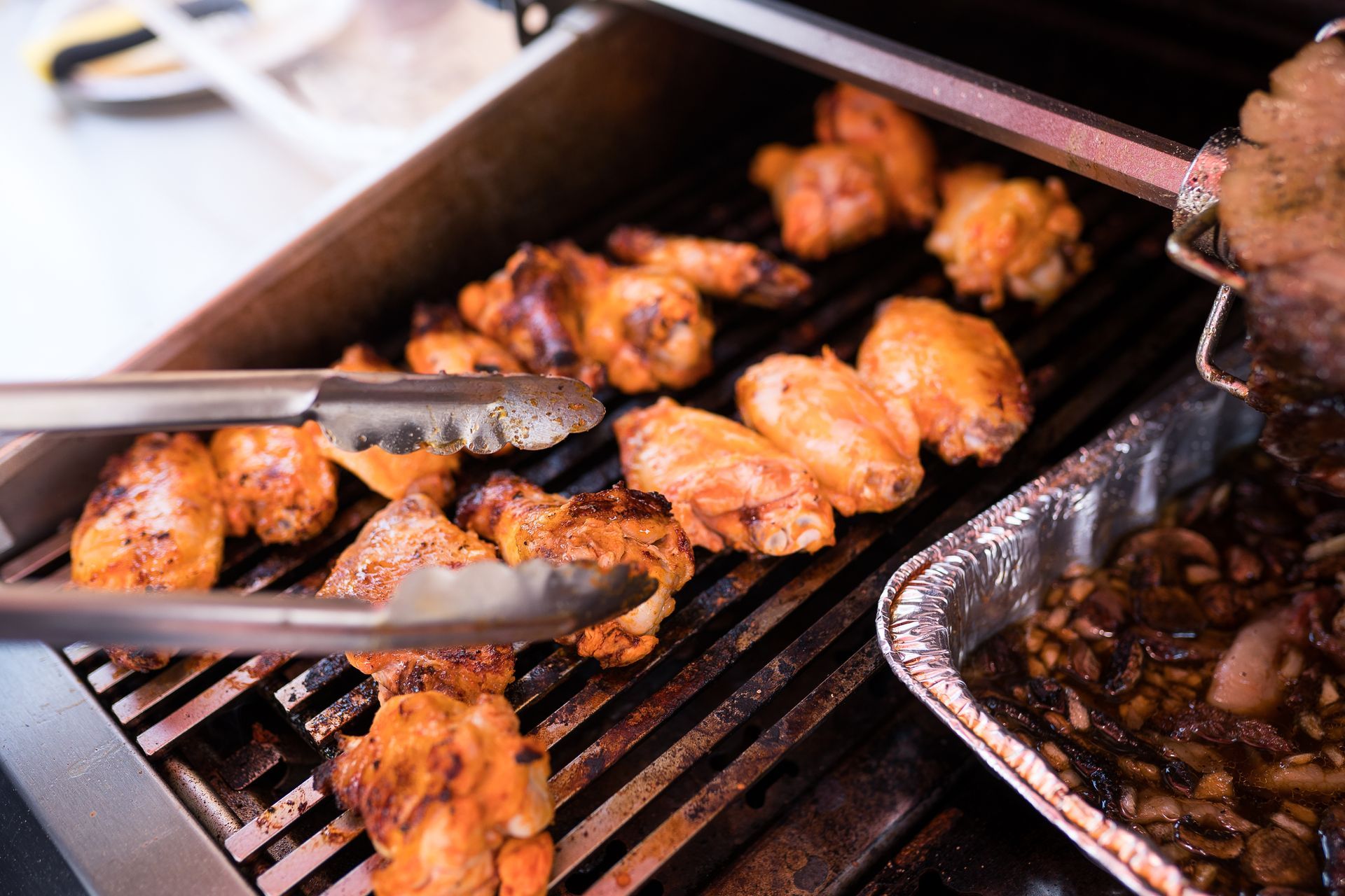 Chicken wings grilling on a BBQ. Metal tongs are turning the wings. A foil pan of food is nearby.