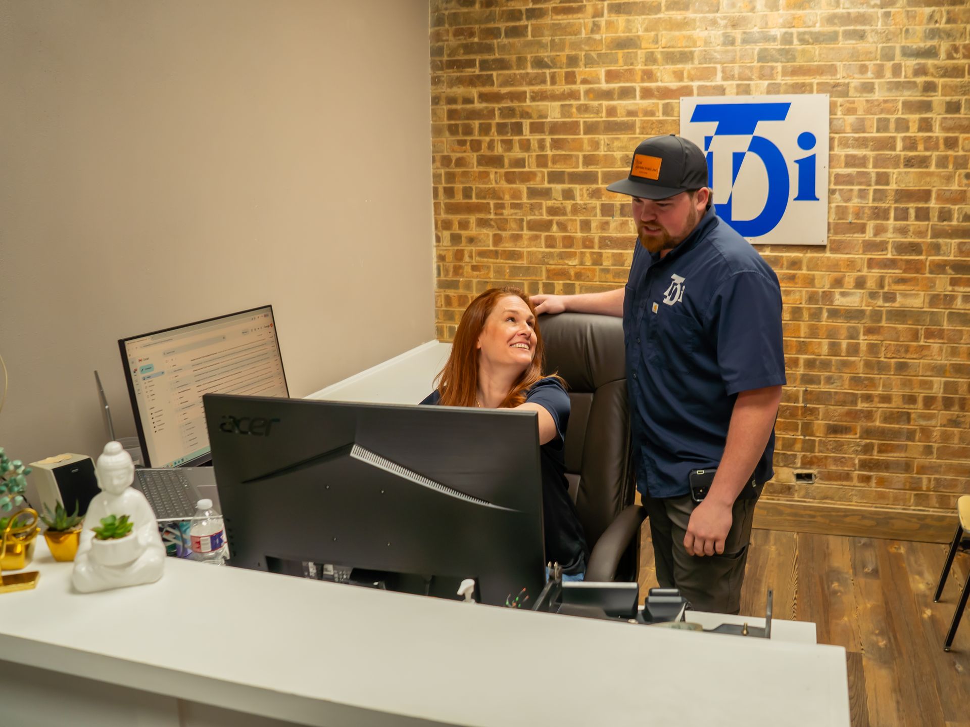 Woman at desk smiles at man, who leans on the chair with a hand on the back, next to a brick wall.