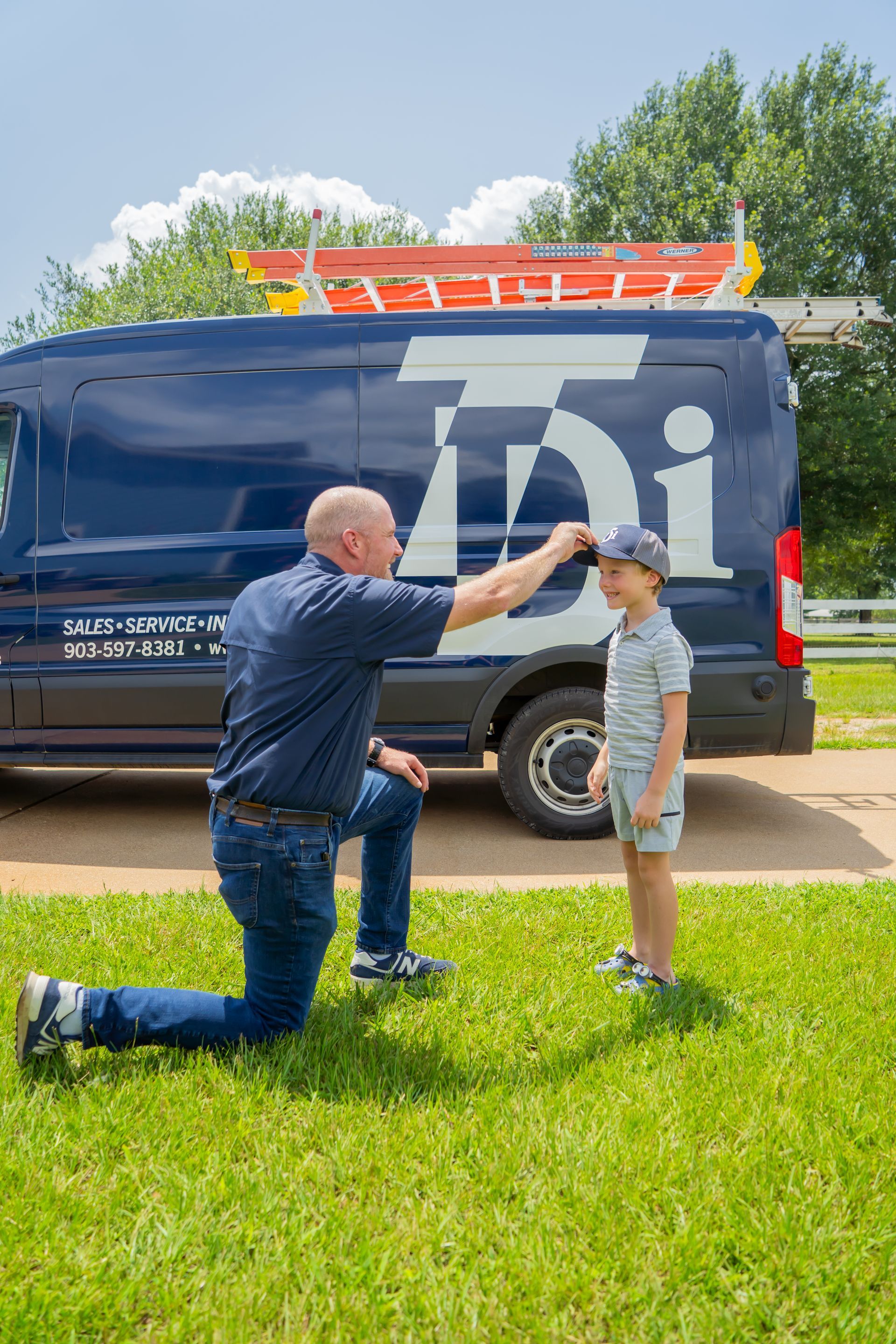 Man kneeling, adjusting a boy's cap, beside a work van, on green grass.