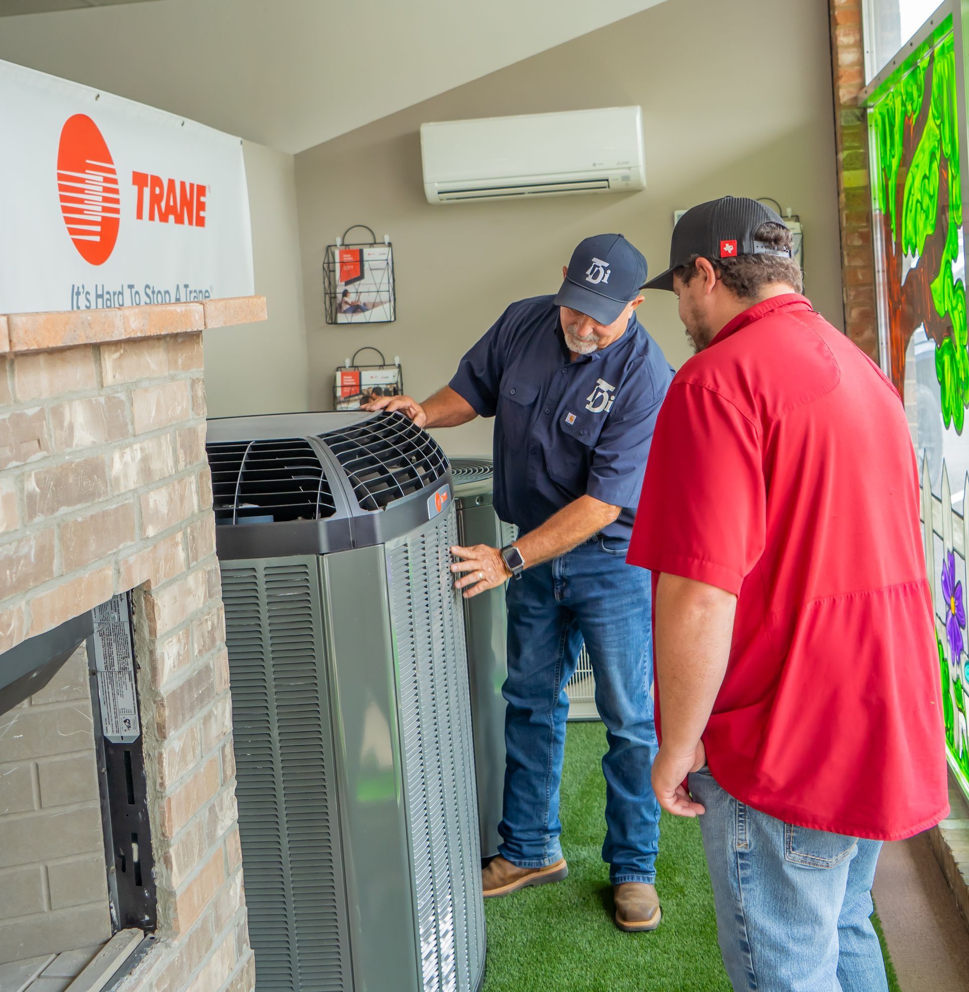 Two men examining a Trane air conditioning unit. One man is wearing a blue shirt, the other a red shirt. Indoor setting.