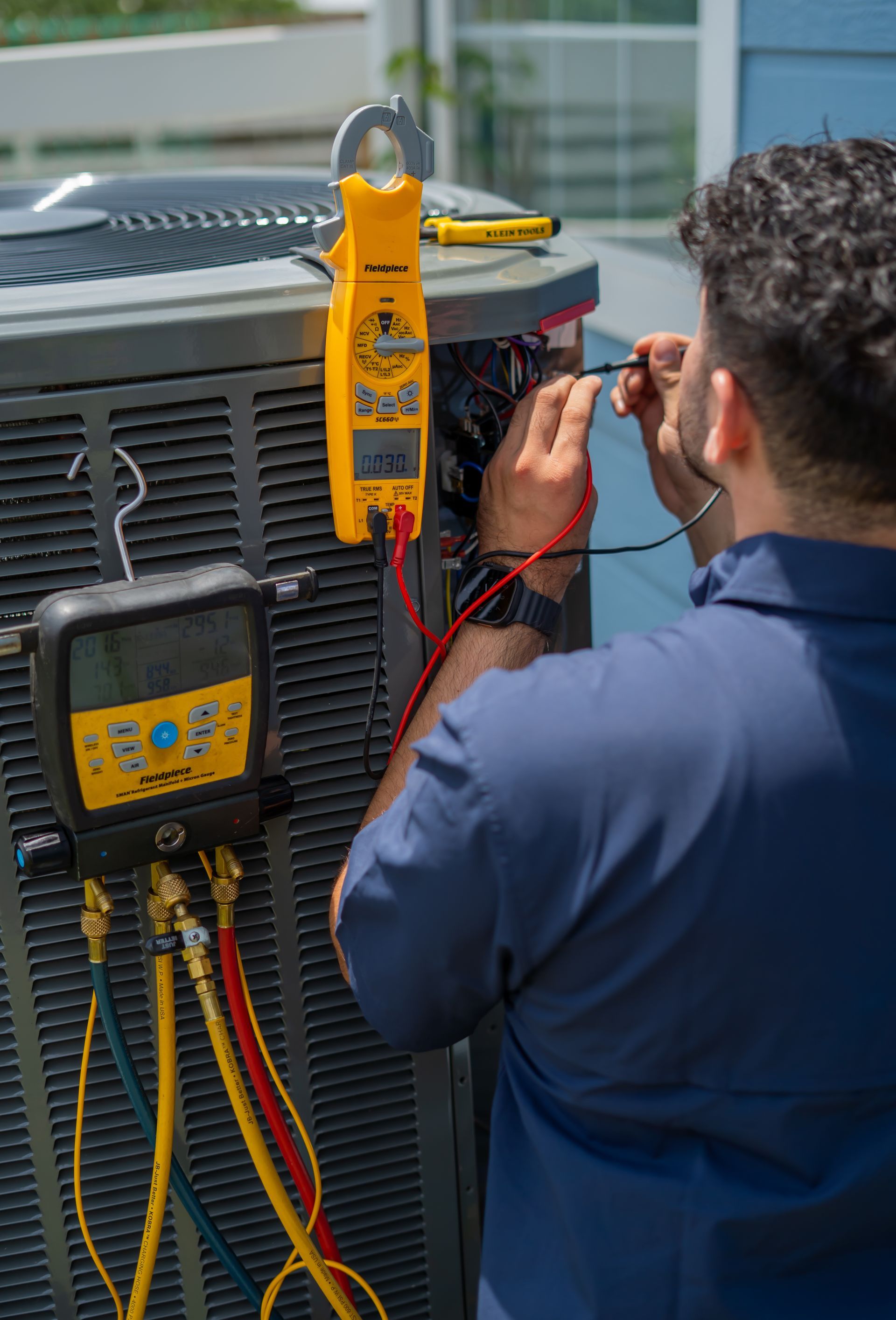 HVAC technician testing an AC unit with yellow and black multimeter. Outdoor setting, sunny.