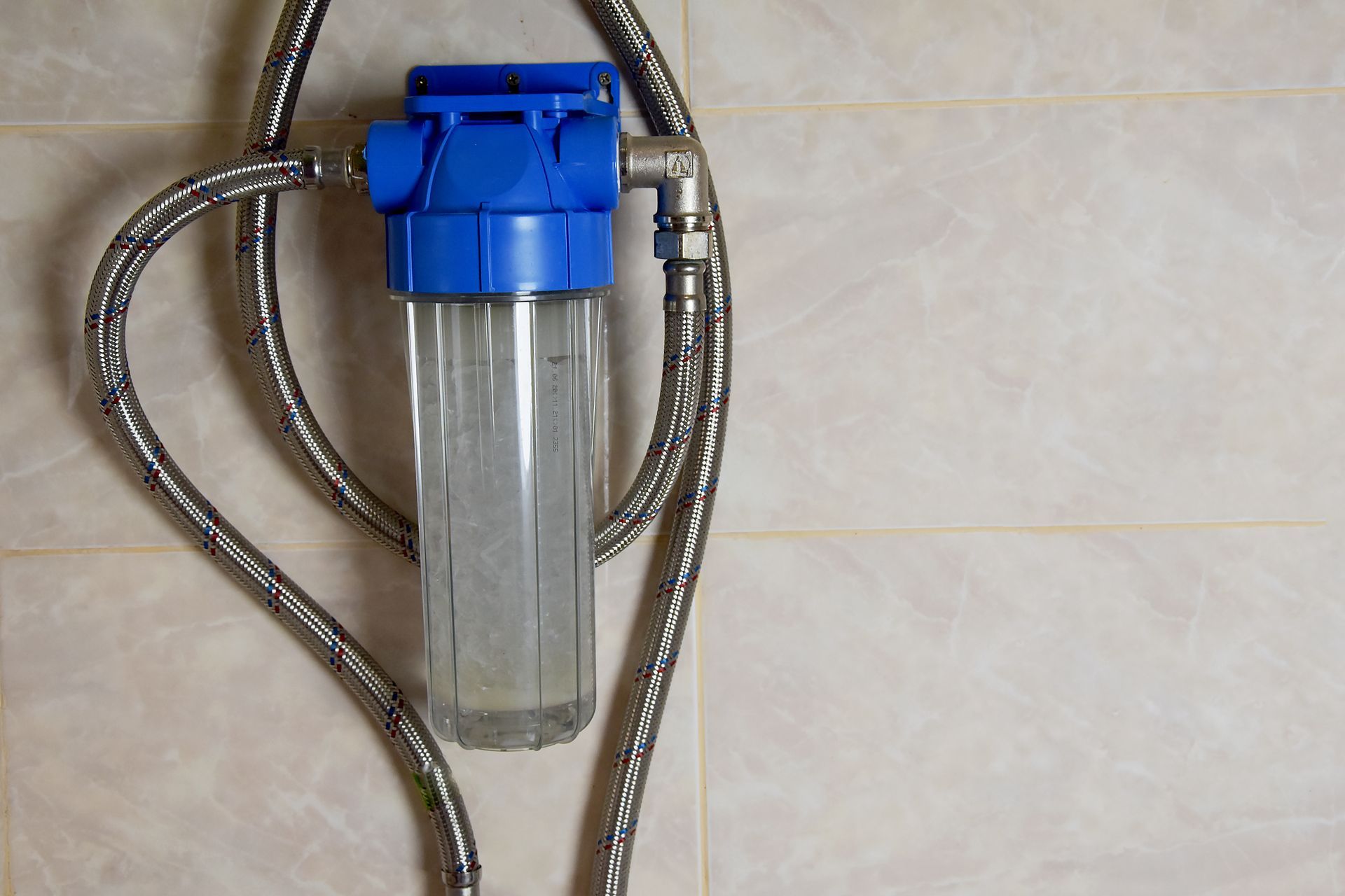 Blue water filter with transparent cylinder, mounted on a tiled wall. Metal hoses connect to the filter.