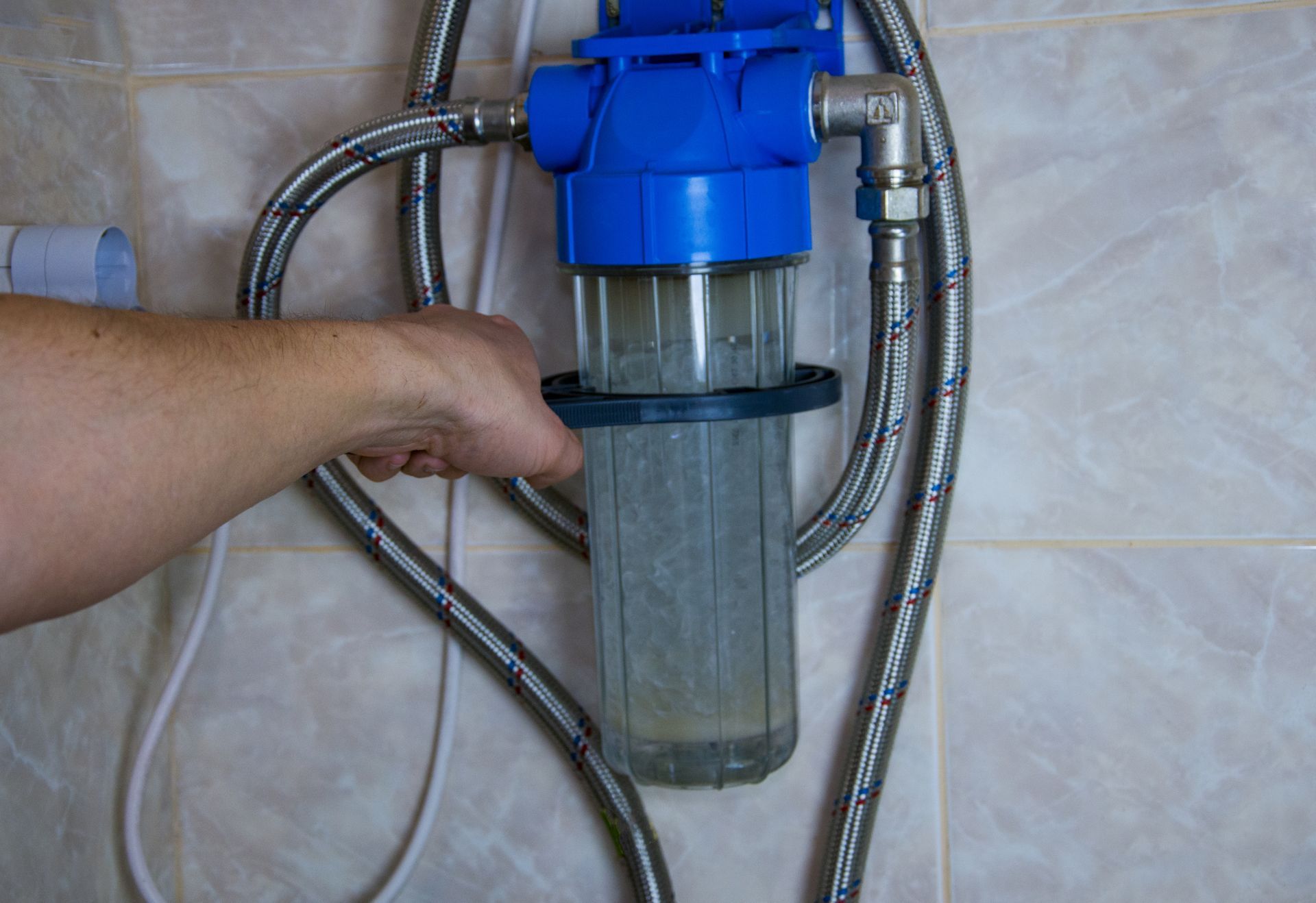 A hand holding a water filter housing. The clear container shows sediment. Blue filter head, metal pipes, and tiled wall.