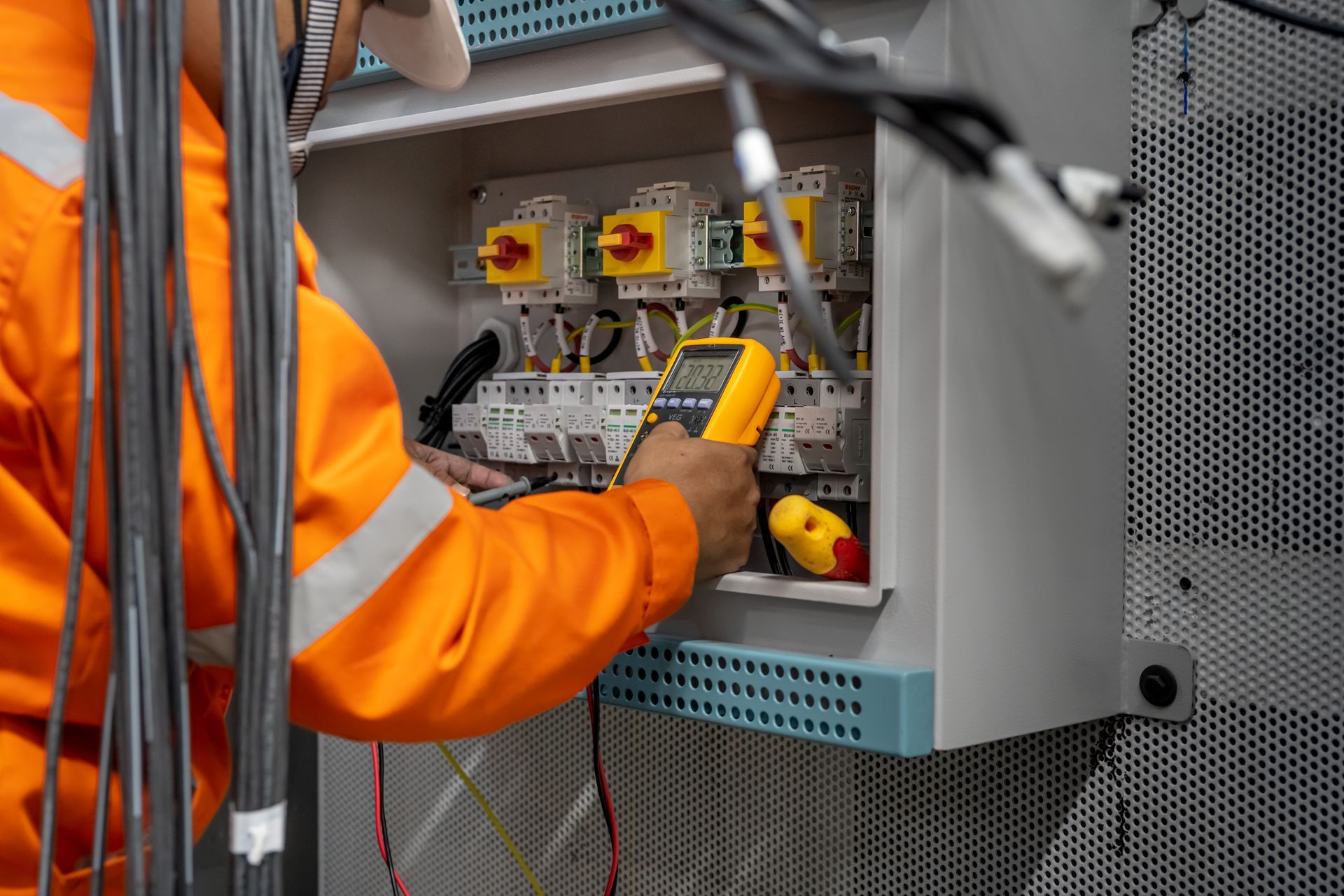 Electrician in orange safety gear using a multimeter on electrical panel.