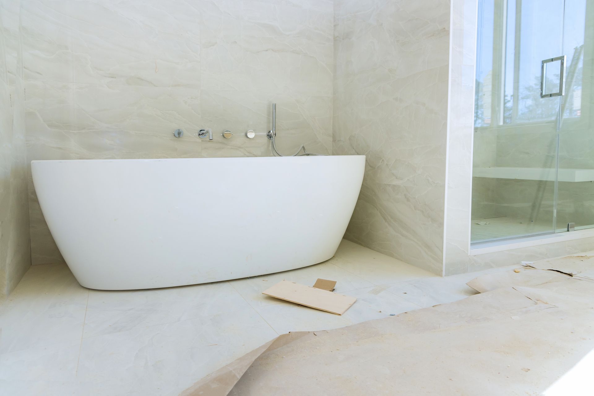 White bathtub in a light-colored bathroom with a glass shower and exposed construction materials.