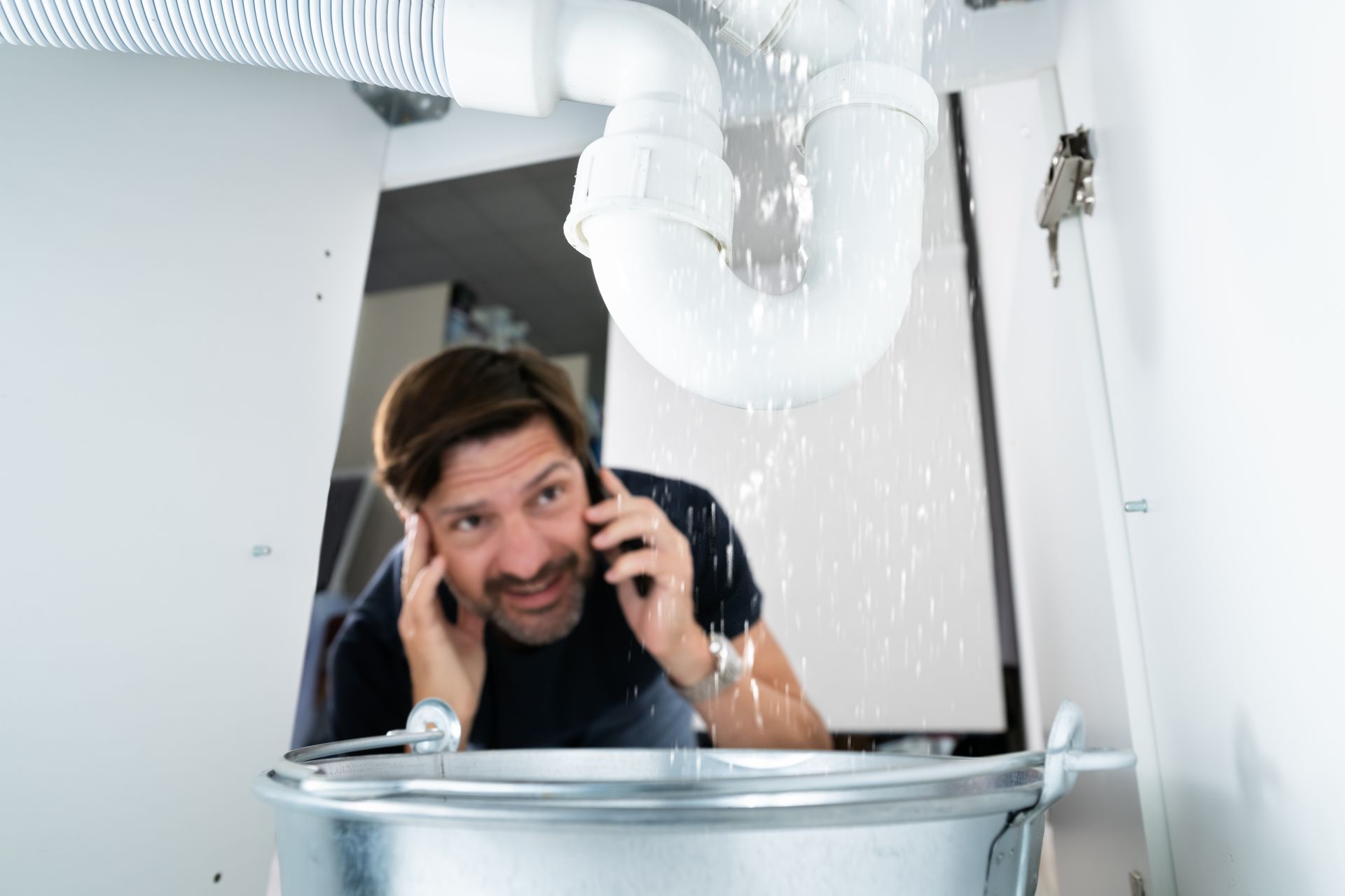 Man on phone, looking concerned at leaky pipe above a metal bucket in a white cabinet. Water is spraying.