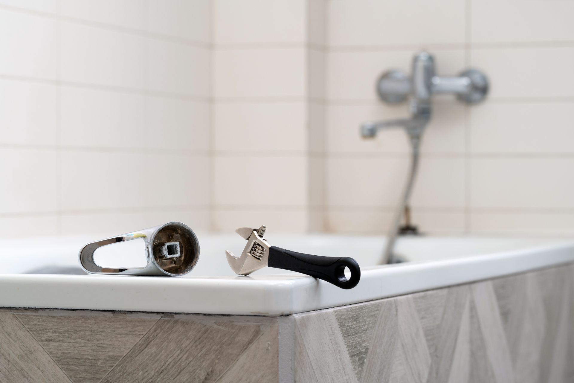 A bathtub with a detached faucet and a repair tool on the edge, against a tiled wall.