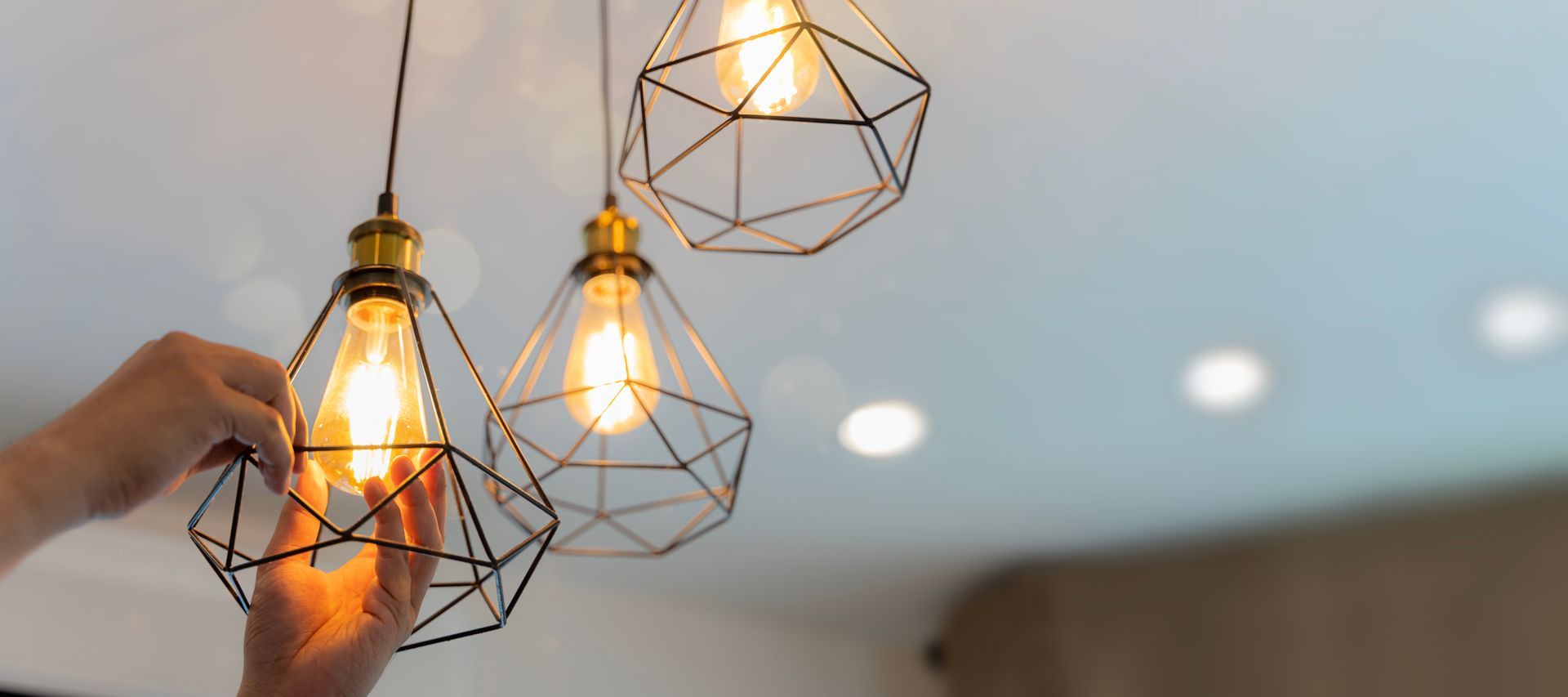 Person's hands adjusting a geometric pendant light fixture with a glowing Edison bulb in a home.