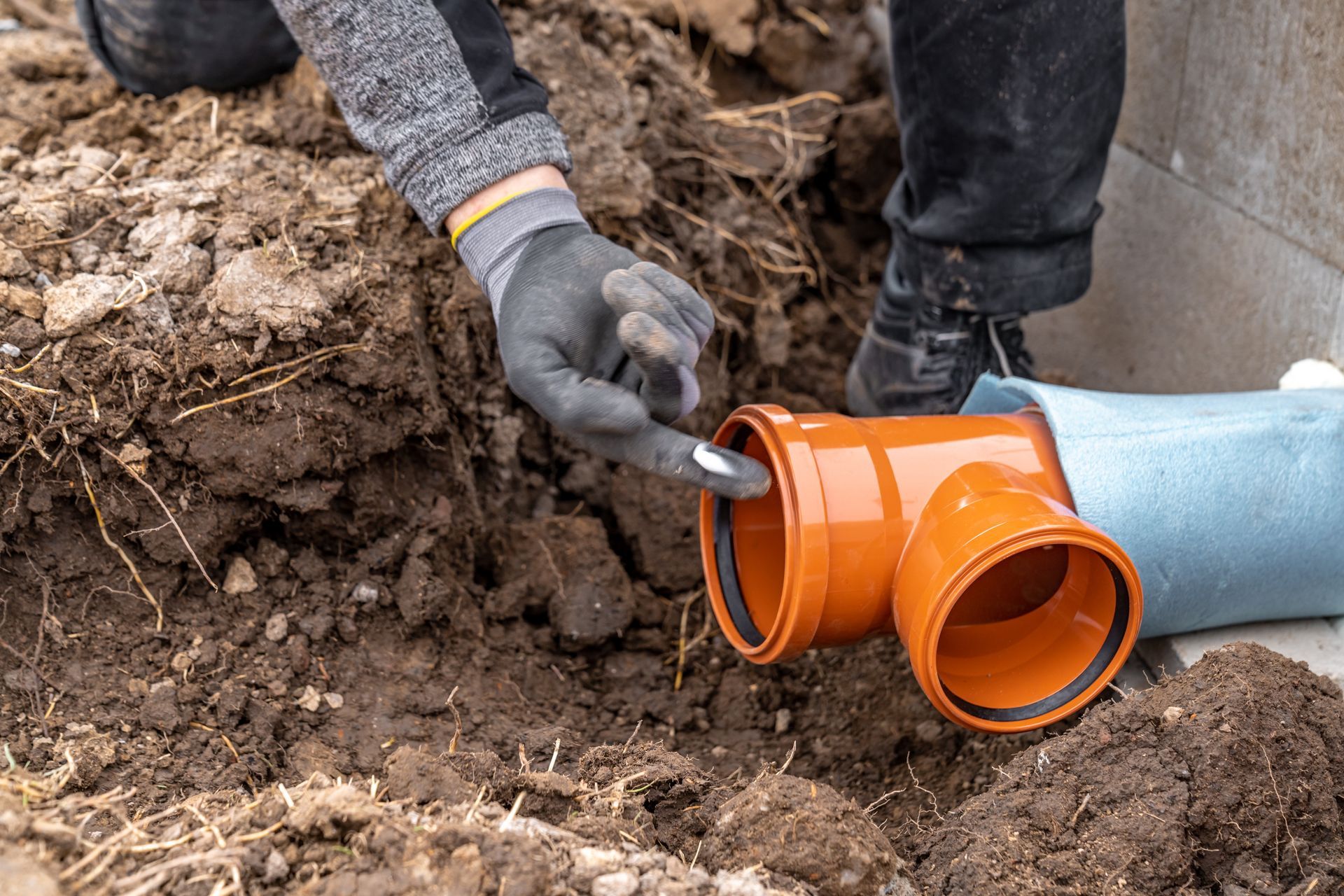Person installing orange plastic plumbing fitting in a trench, working outside.