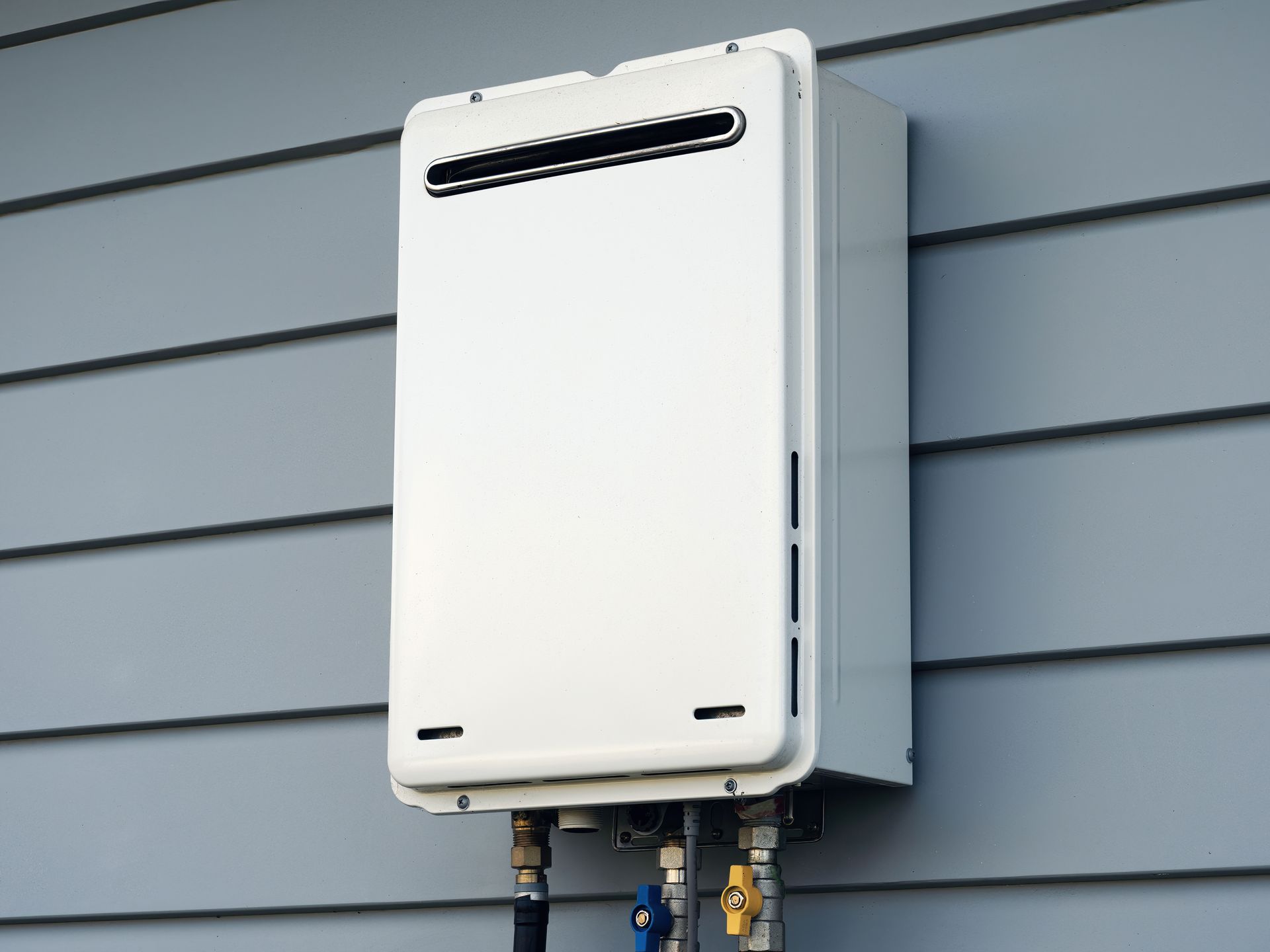 White tankless water heater mounted on a gray exterior wall with blue and yellow valves.