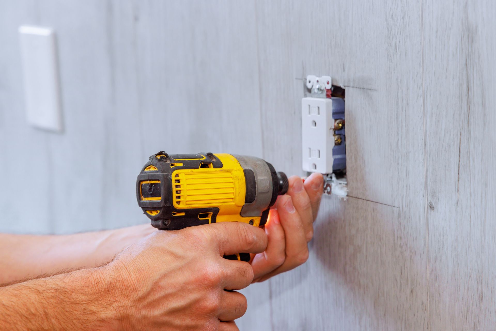 A person using a yellow and black power drill to install an electrical outlet on a light gray wall.