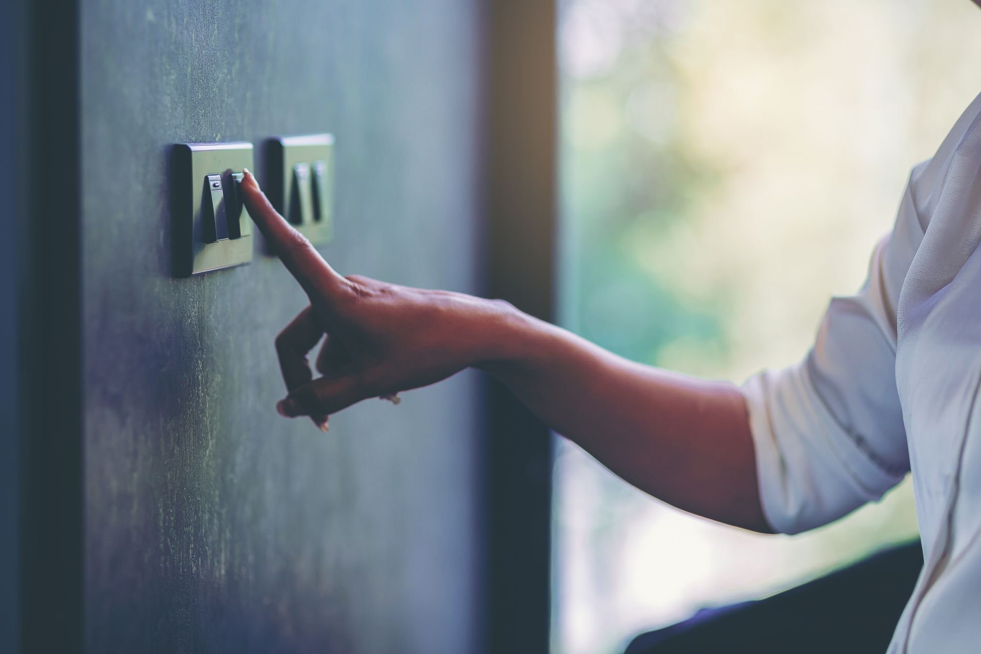 Person's hand presses a light switch on a dark wall. Sunlight streams through a window.