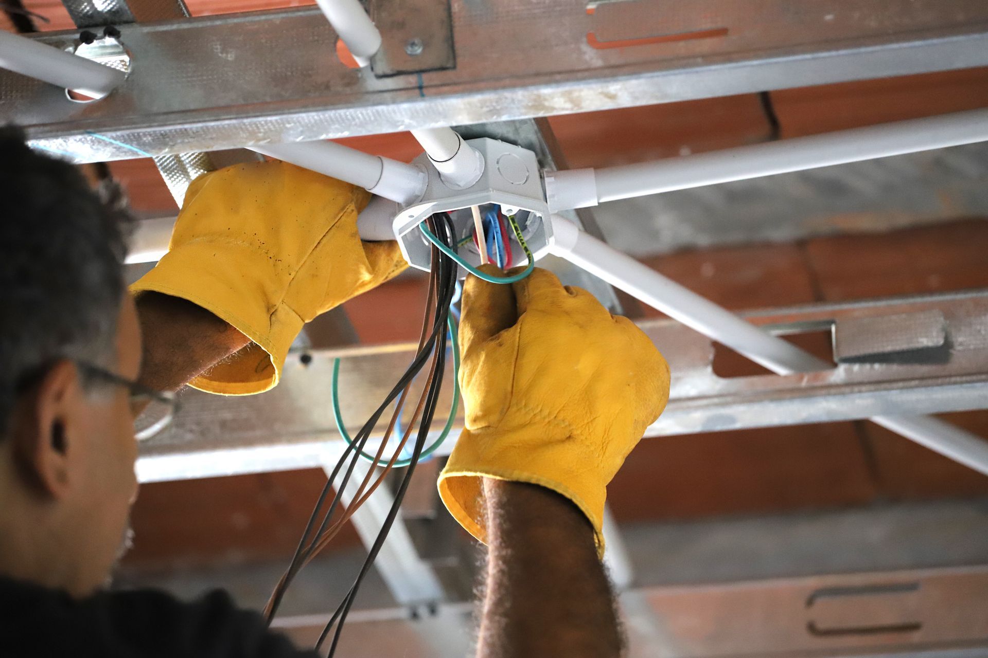 Electrician working on wiring inside a ceiling junction box, wearing yellow gloves.