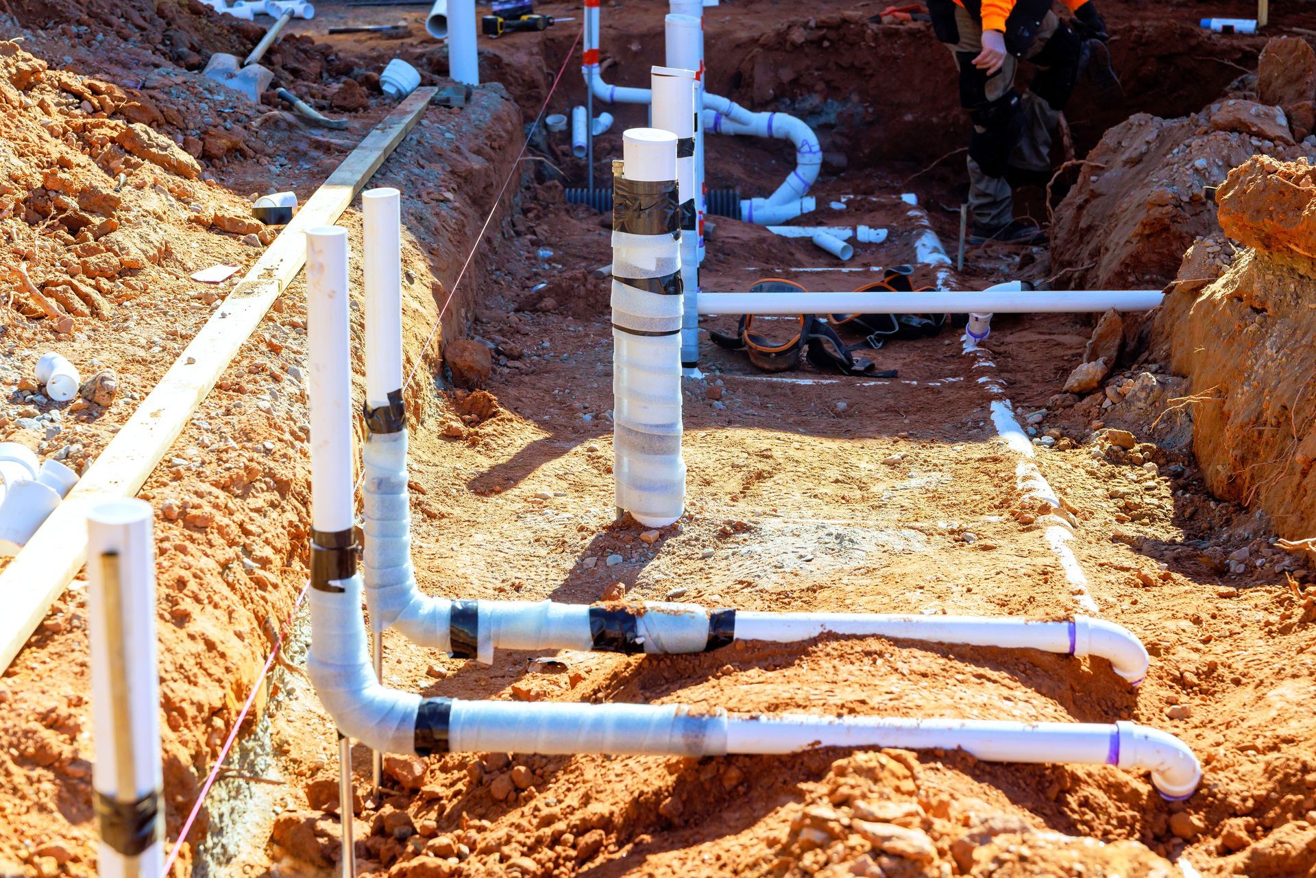 Underground PVC plumbing installation in a dirt trench; white pipes, black tape, red-brown soil.
