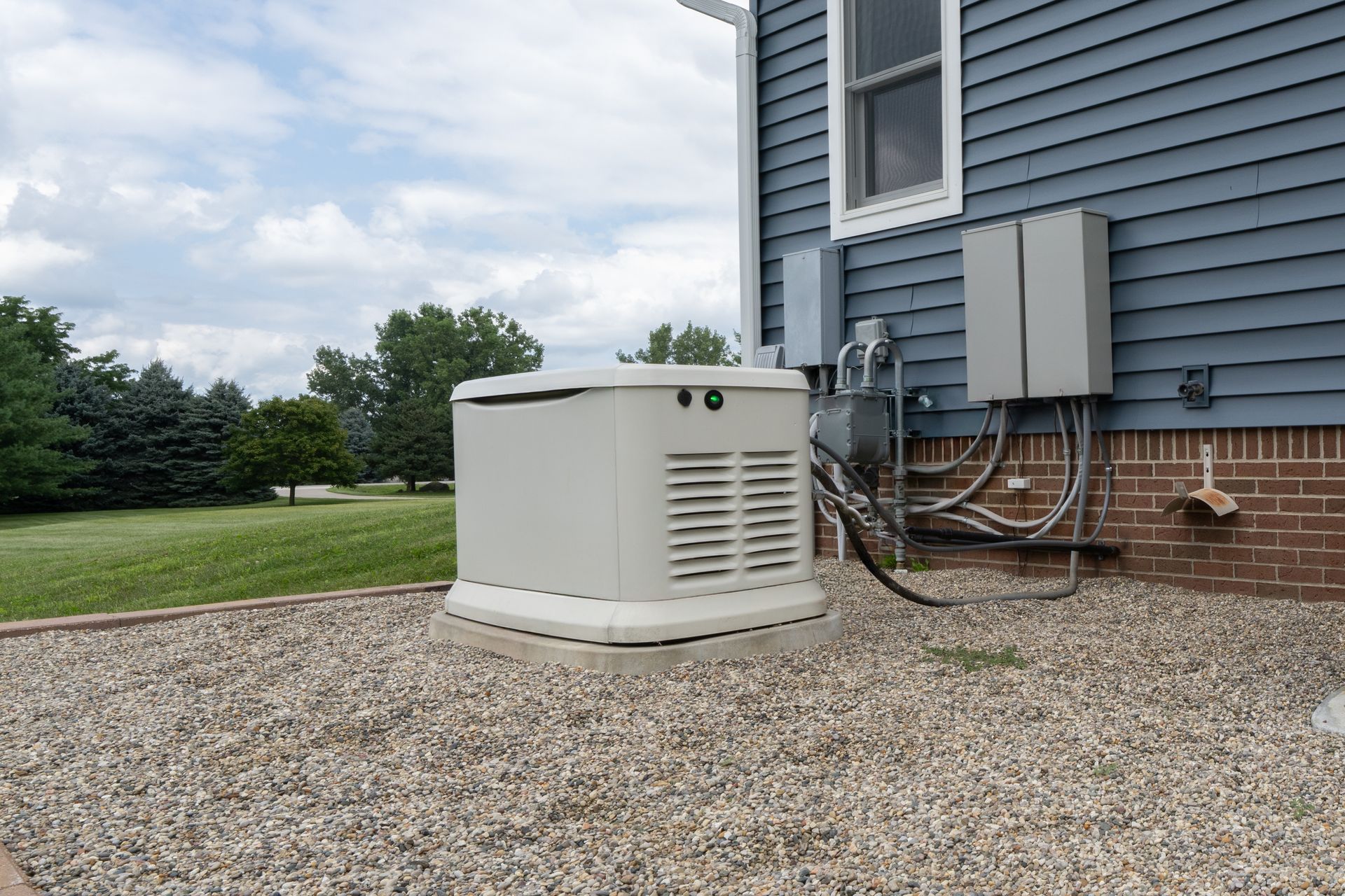 A standby generator on a gravel base next to a blue-sided house with electrical components.