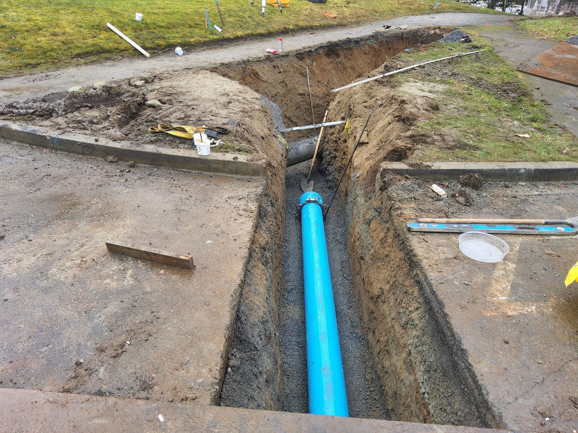 Trench dug in concrete, blue pipe installed. Construction site with tools and markings on grass and sidewalk.