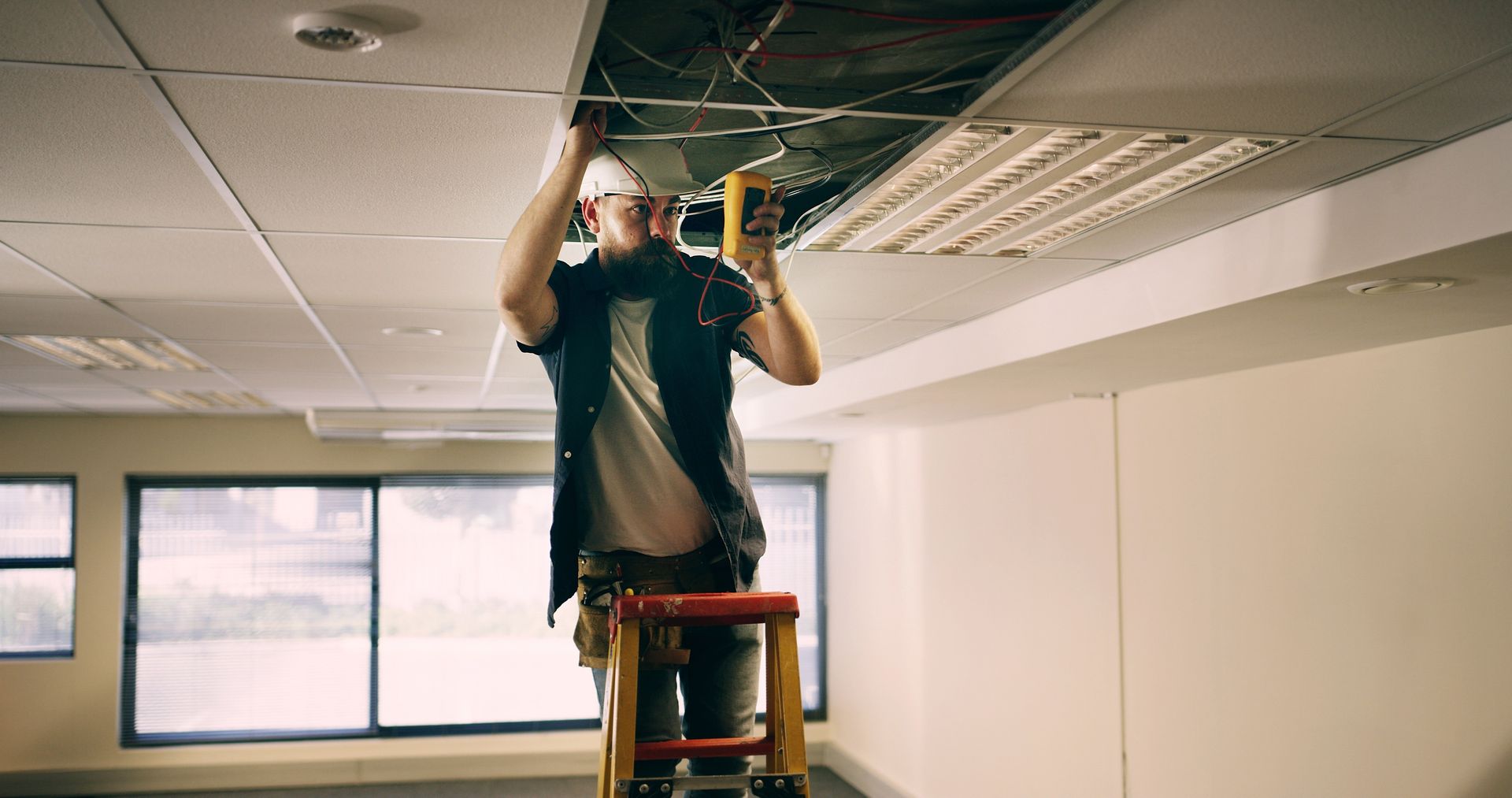 Electrician on ladder working on ceiling wiring in a room with windows and white walls.