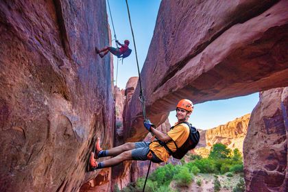 A man and woman are rappelling under a stone arch with a canyon below.