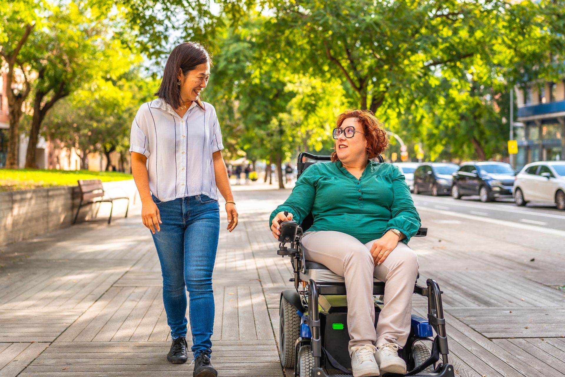 Woman in wheelchair on sidewalk converses with woman walking beside her in front of a city street.