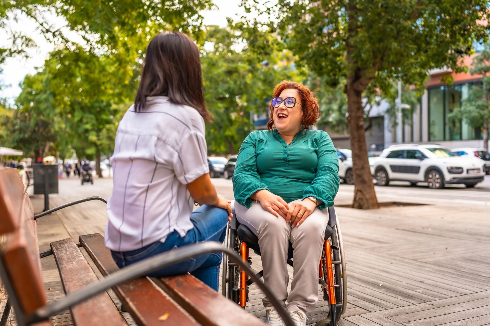Two women talking on a bench outdoors; one in a wheelchair, smiling.
