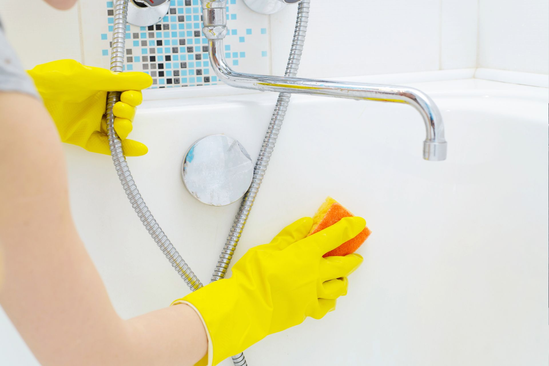 Person in yellow gloves cleaning a white bathtub with an orange sponge.