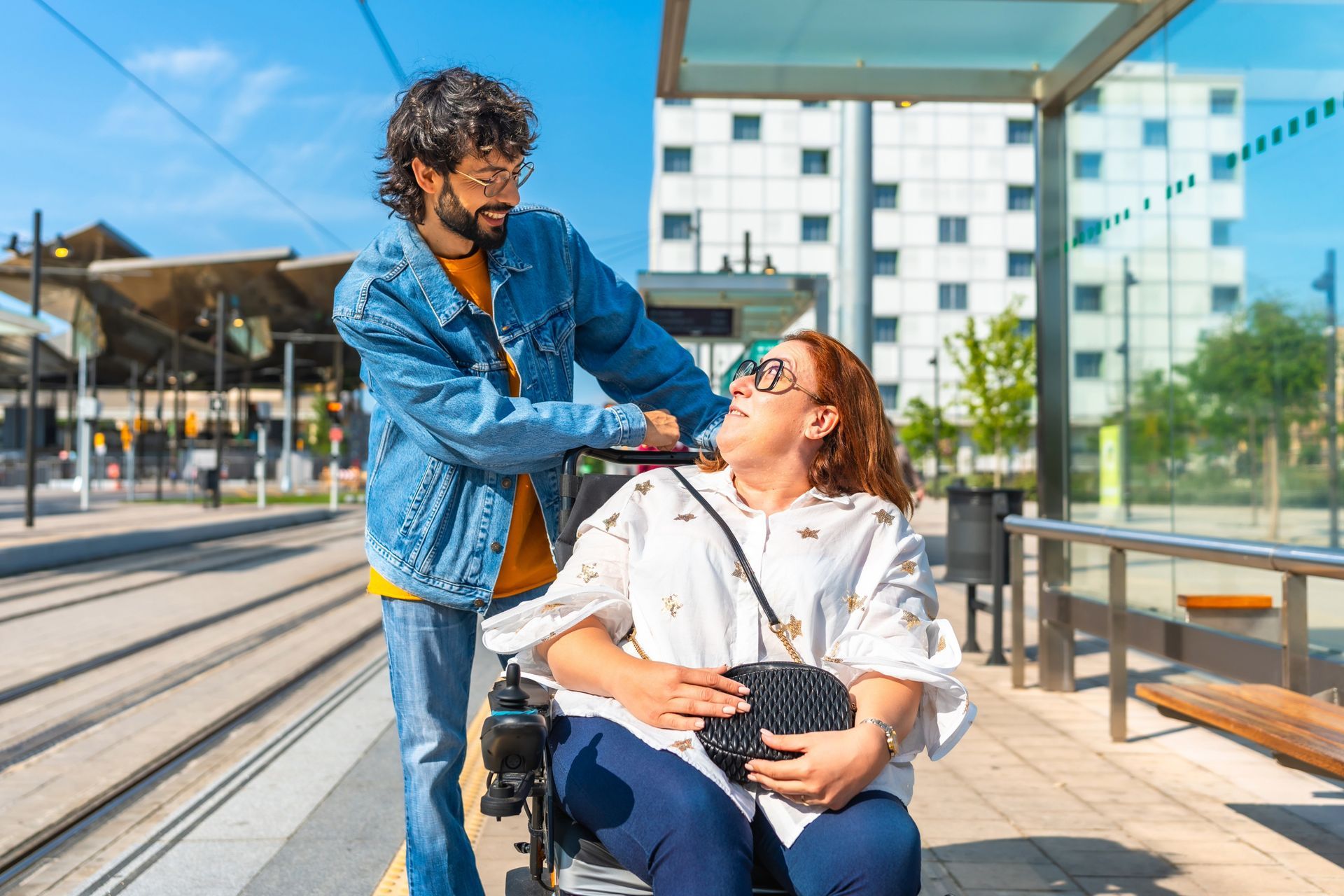 Man assisting person in wheelchair on a train platform. They're smiling, near tracks, a building in background.