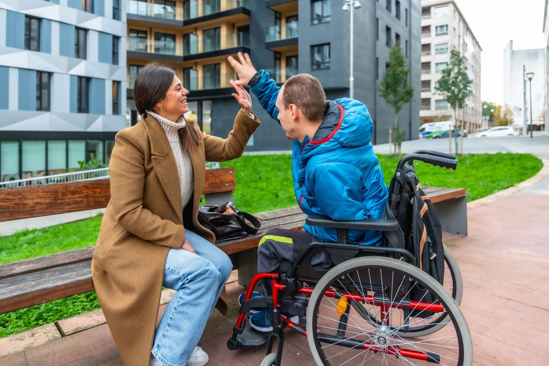 Woman in brown coat high-fives a person in a wheelchair in front of buildings and greenery.