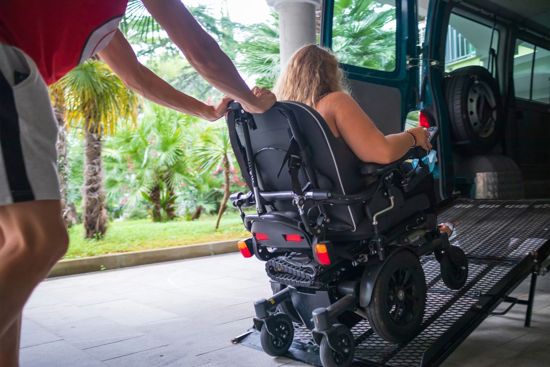 Person assisting someone in a wheelchair onto a vehicle using a ramp outdoors.