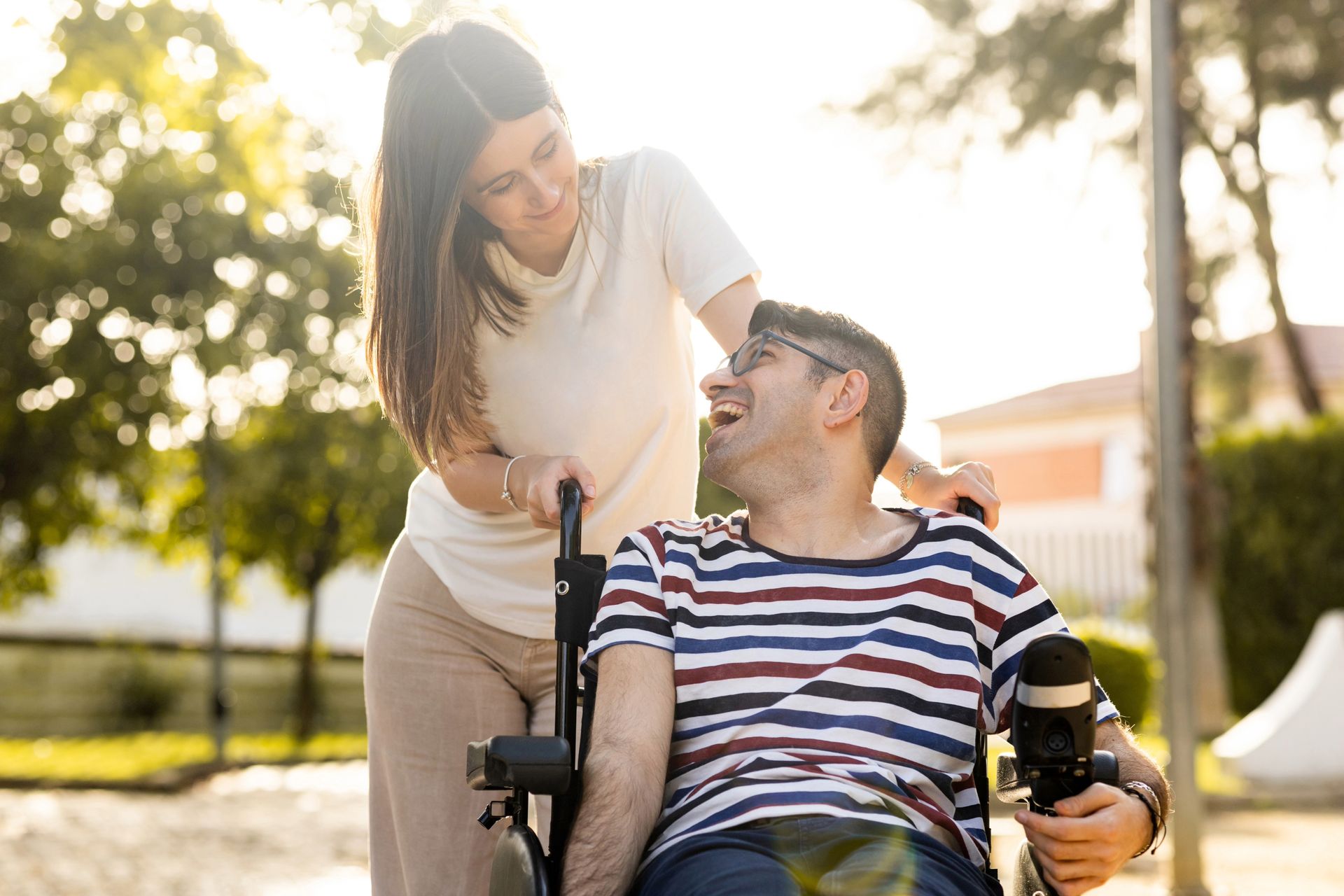 Woman pushing a man in a wheelchair; both smile. Sunny outdoor setting. Man holds a camera.