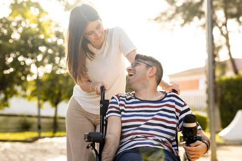 Woman pushing a man in a wheelchair; both smile. Sunny outdoor setting. Man holds a camera.