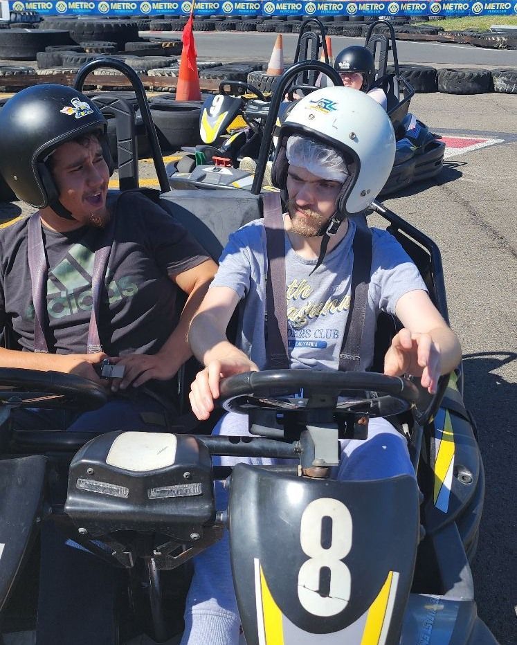 Two people wearing helmets in a go-kart, smiling. Number 8 visible on the kart. Outdoor setting with track.