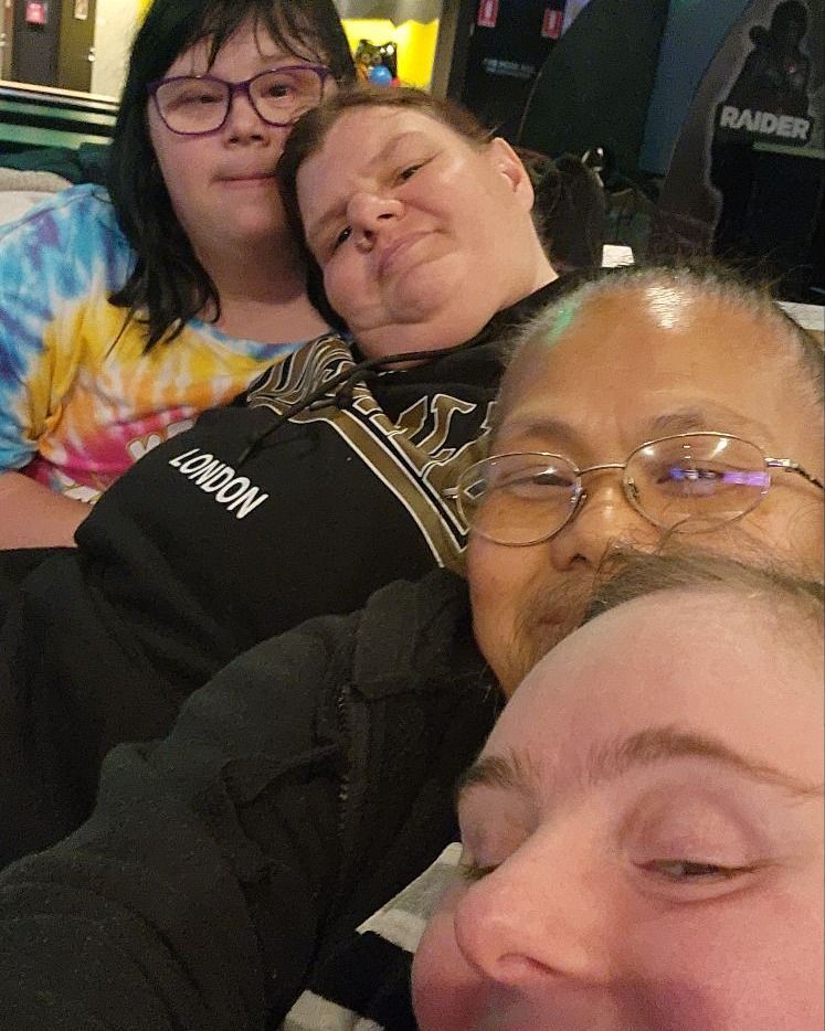 Four people smiling together indoors. Two wearing glasses, one in tie-dye.